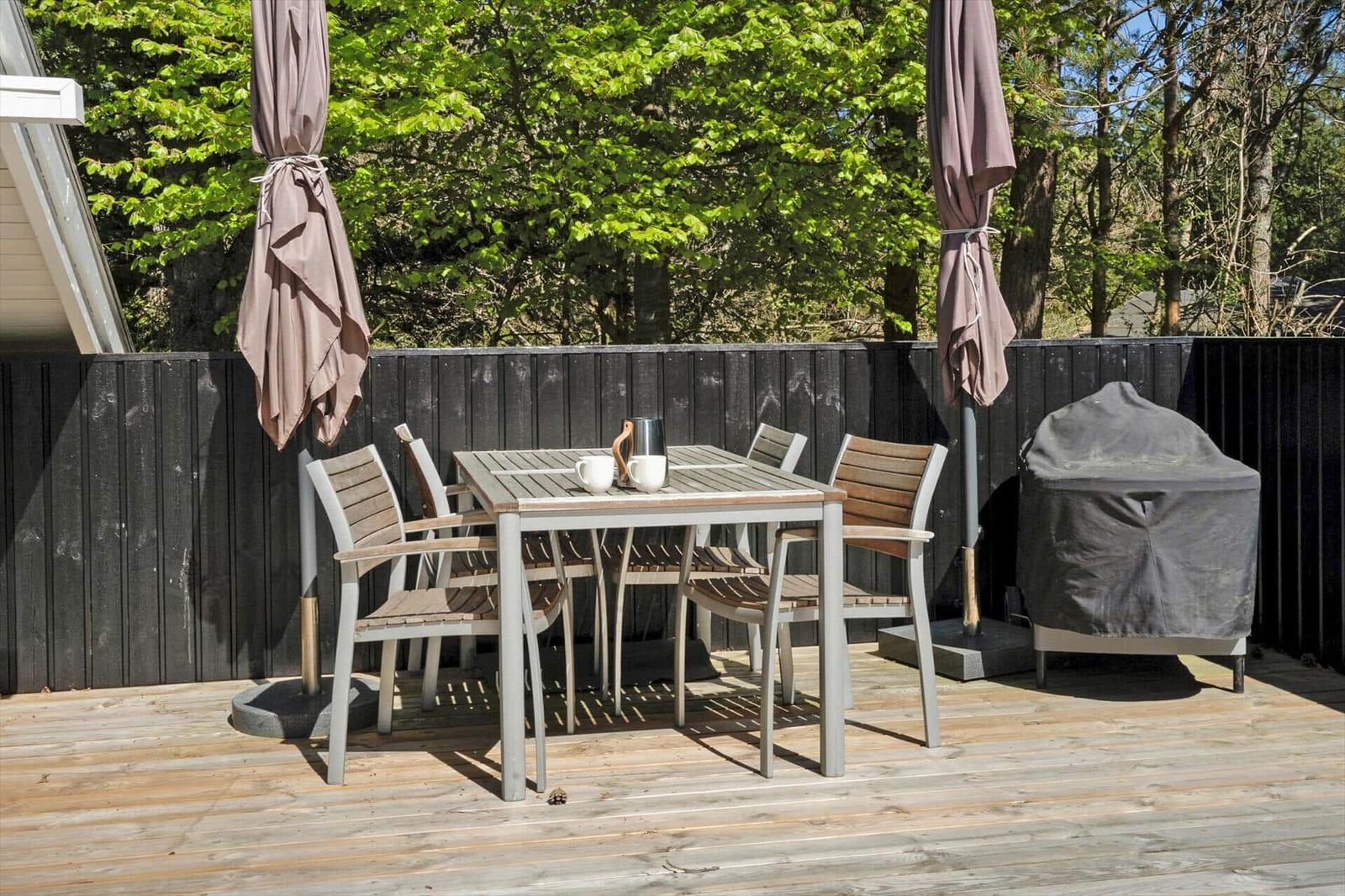 Terrasse mit Tisch, Stühlen und zwei geschlossenen Sonnenschirmen. Grill ist mit Abdeckung versehen.
