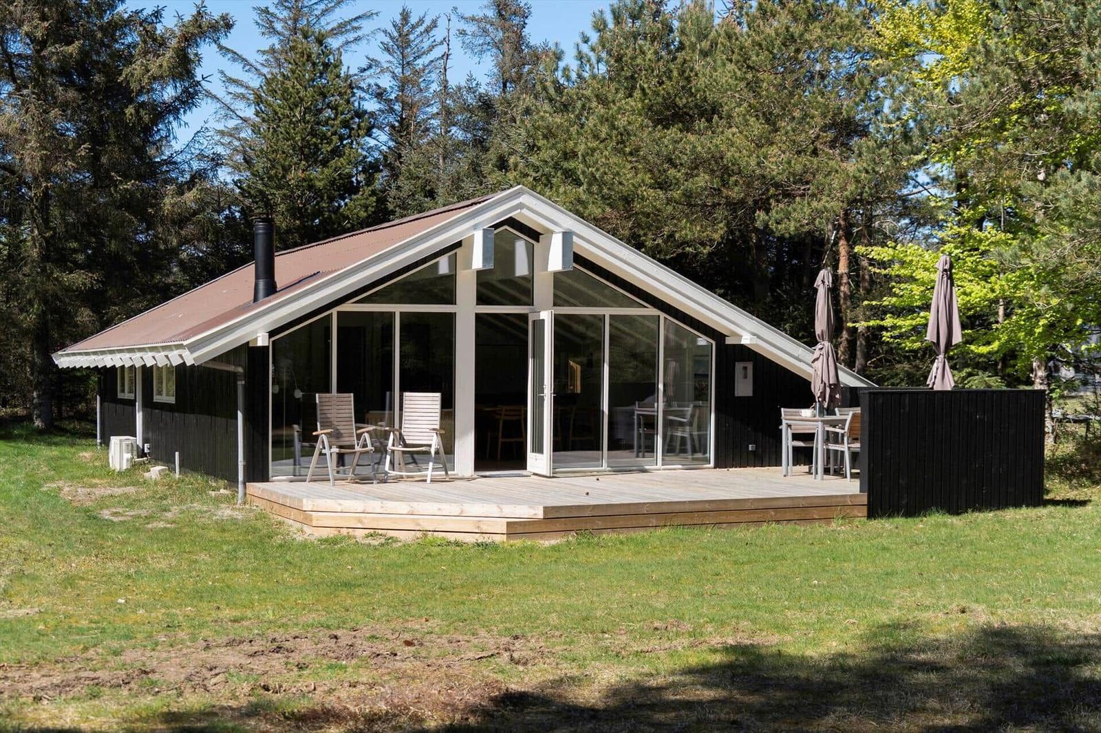 Moderne Villa mit großem Holzterrasse und Glasfront. Umgeben von Wald und Gras.
