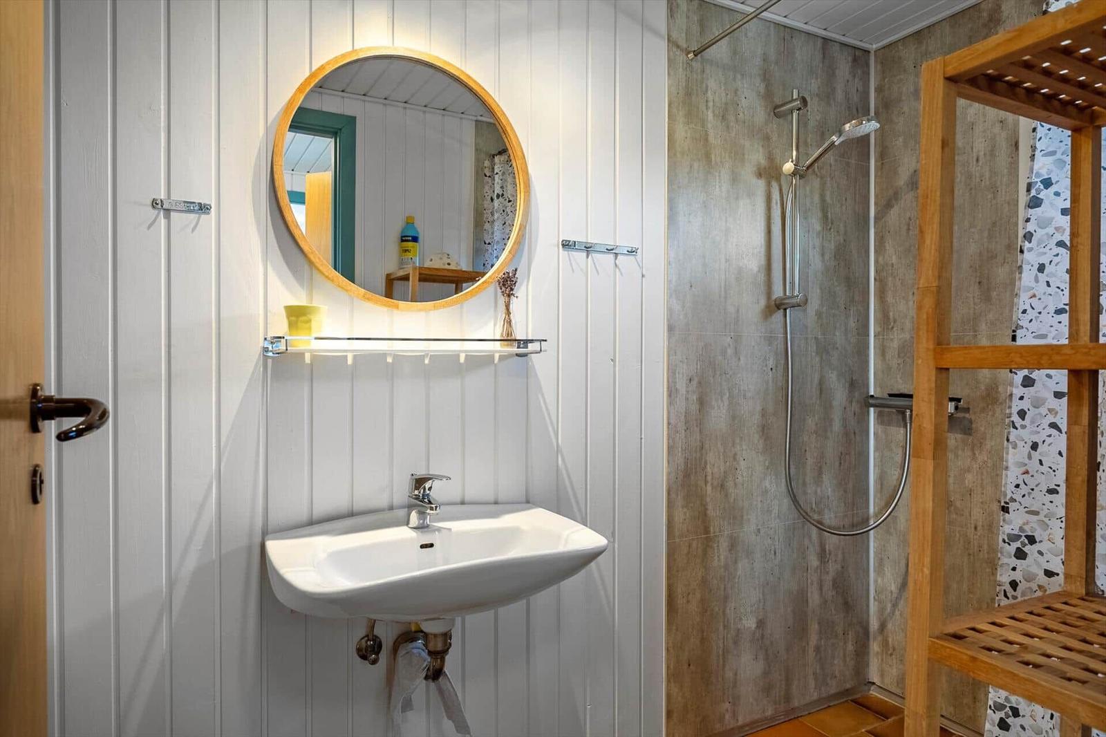Bathroom with sink, round mirror, and shower with wooden shelf.