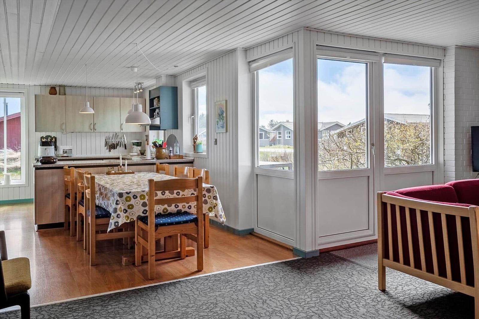 Dining room with table, chairs, and kitchen area. Large windows with view outside.