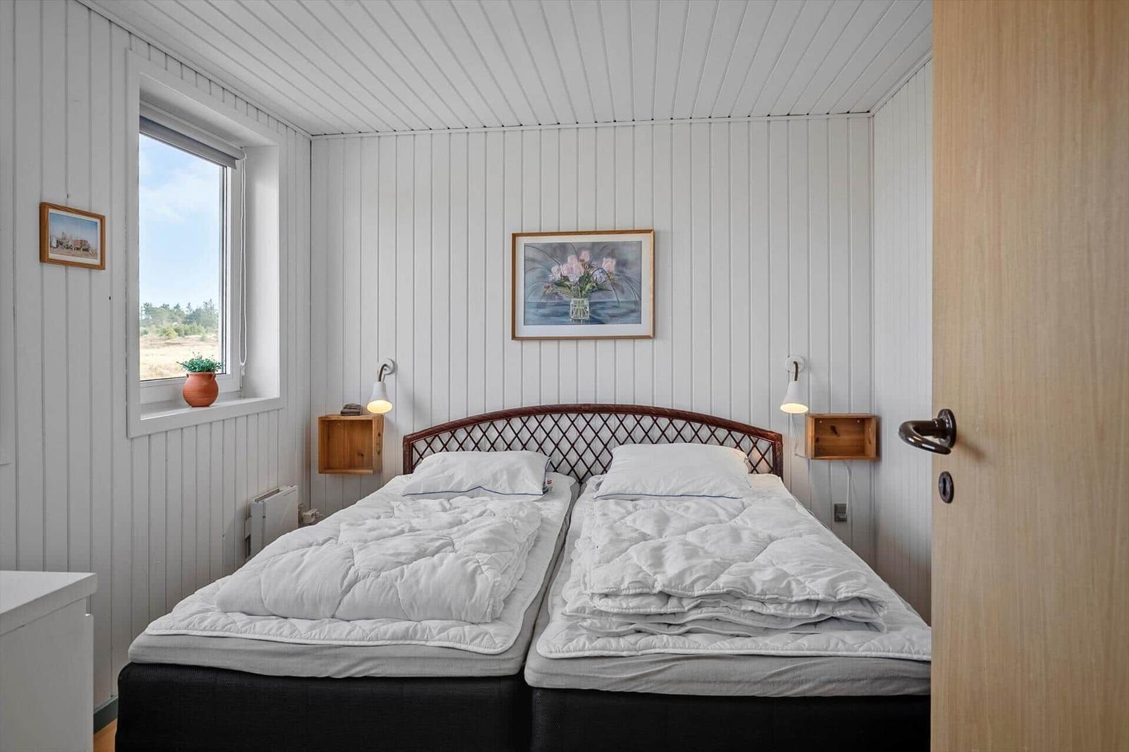 Two beds with white covers, window with view of field, wall pictures and wooden shelves.