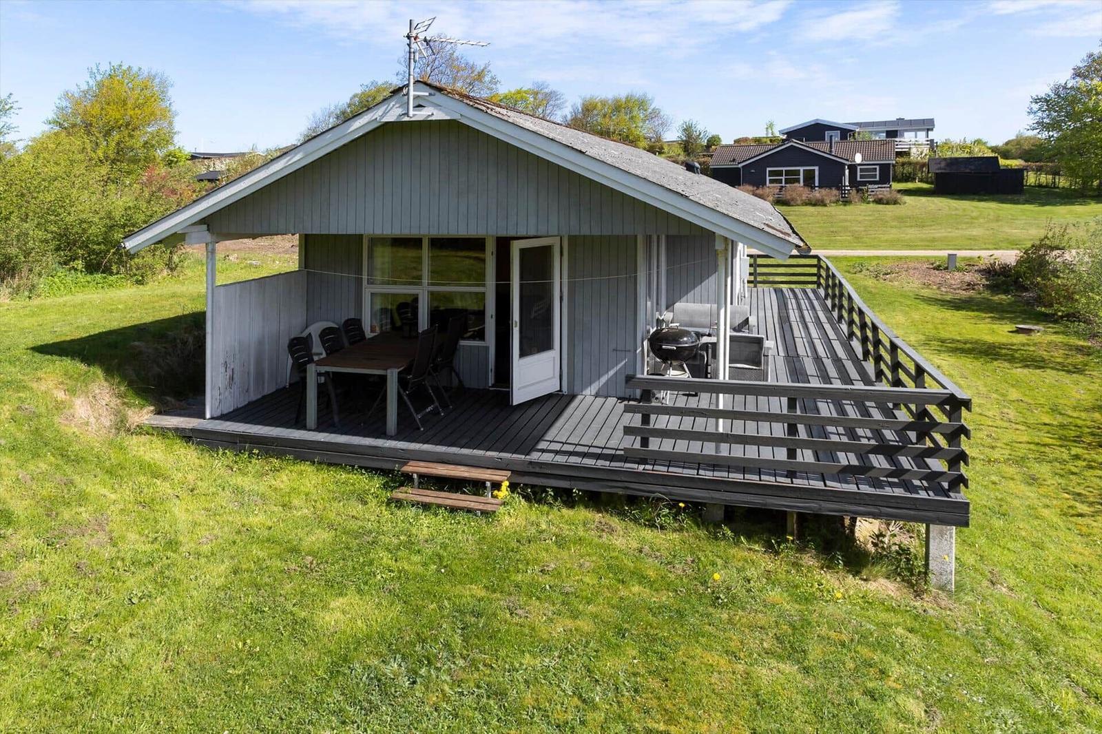 Holzveranda mit Grill und Tisch. Haus mit Dach und Antenne. Grünfläche im Hintergrund.