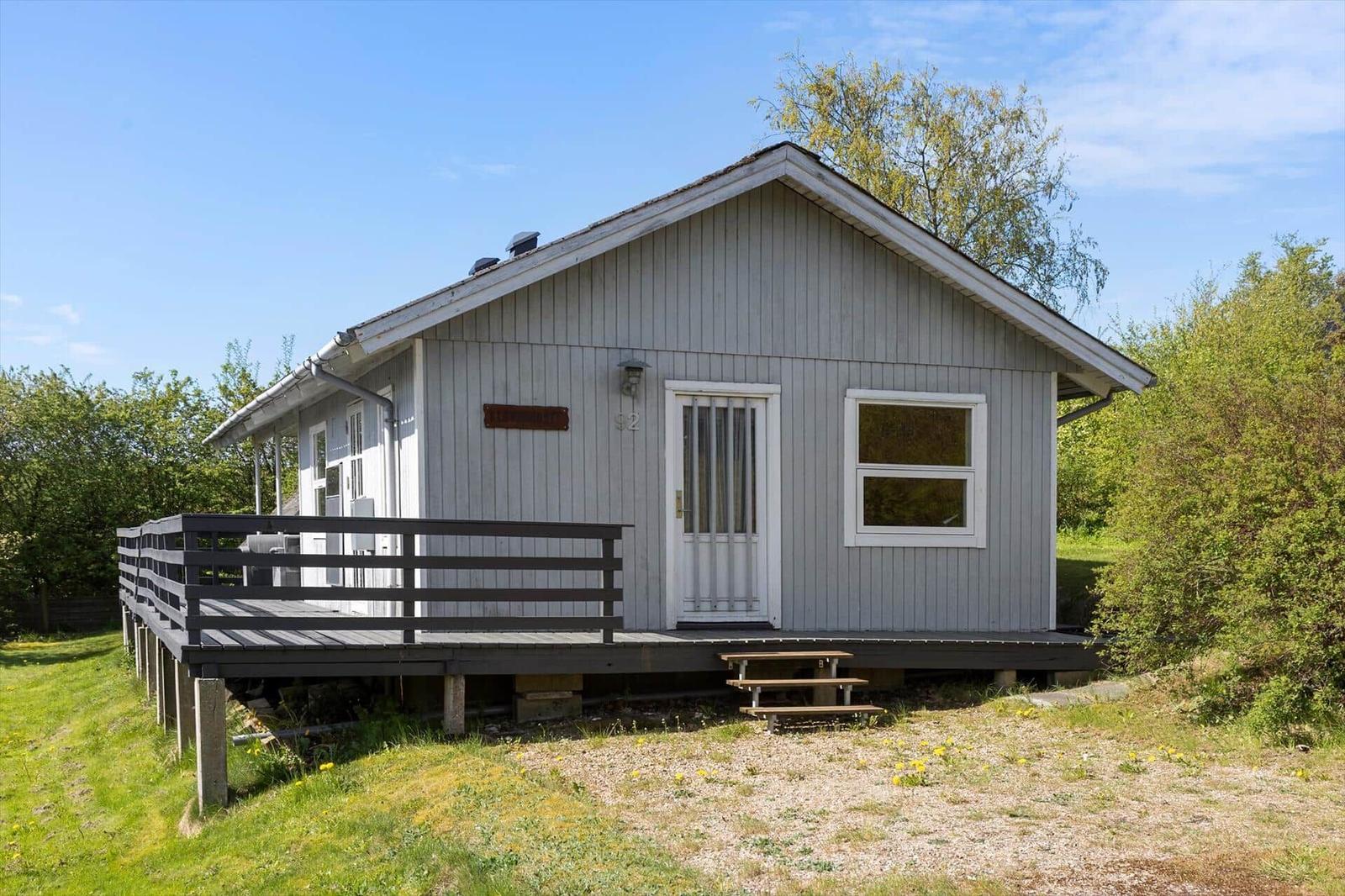 Holzhütte mit Terrasse und Garten. Tür und Fenster sind weiß. Umgeben von Bäumen.