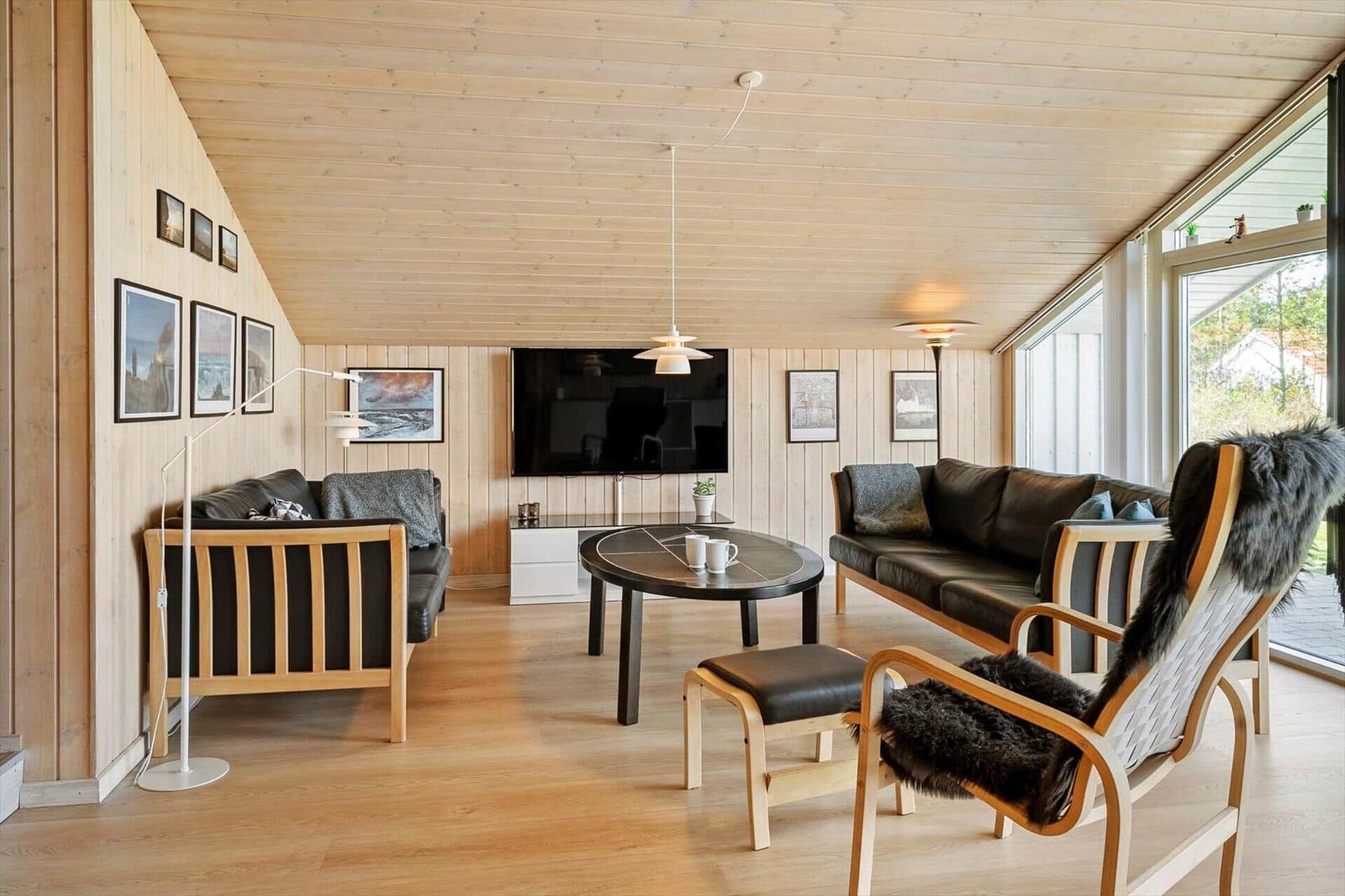 Wohnzimmer mit Holzwänden, Sofa, Sessel und Fernseher. Große Fenster bieten viel Licht.