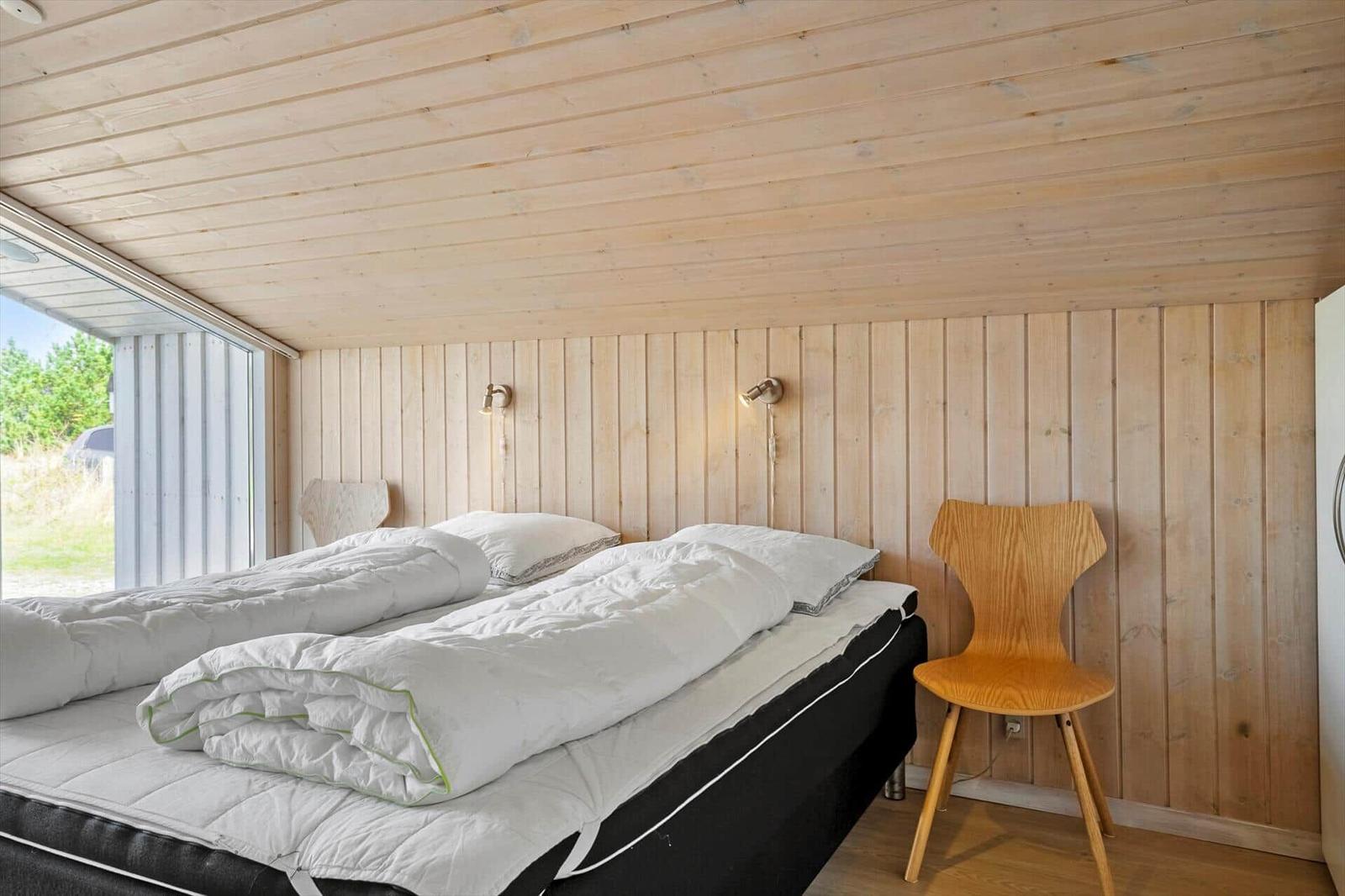 Schlafzimmer mit Holzwand, Bett und Stuhl. Fensterblick auf Außenbereich.