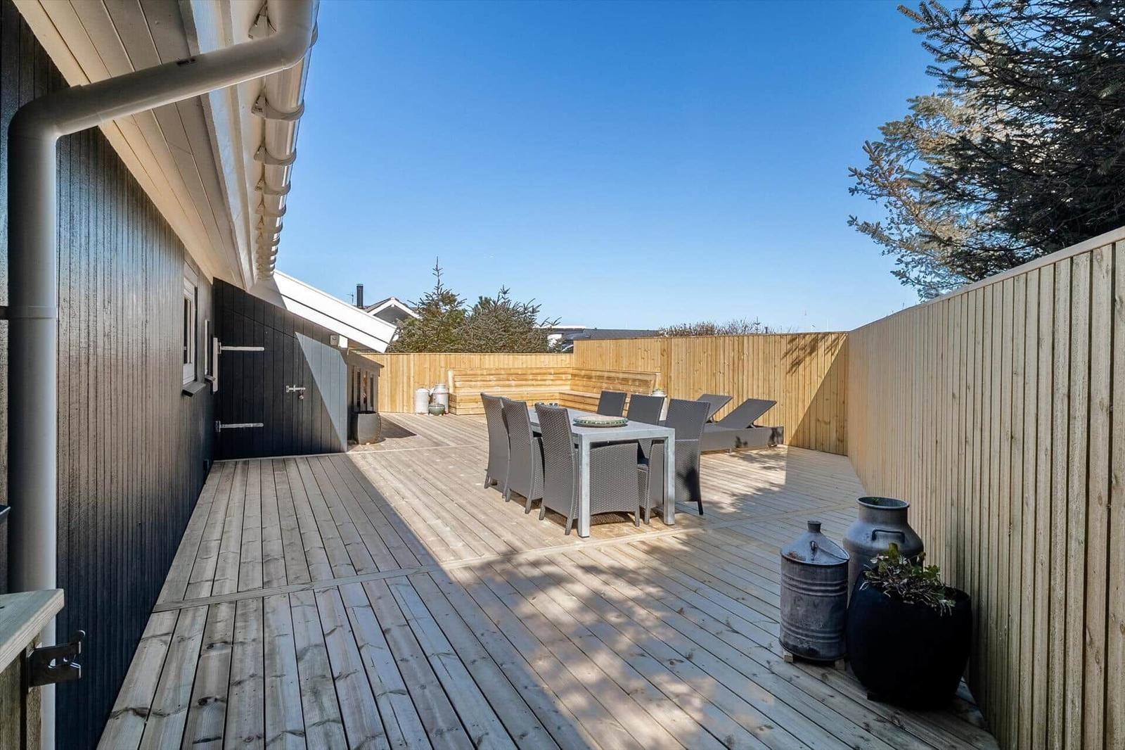 Holzterrasse mit Tisch, Stühlen und Liegen. Umgeben von Holzzaun und Bäumen.