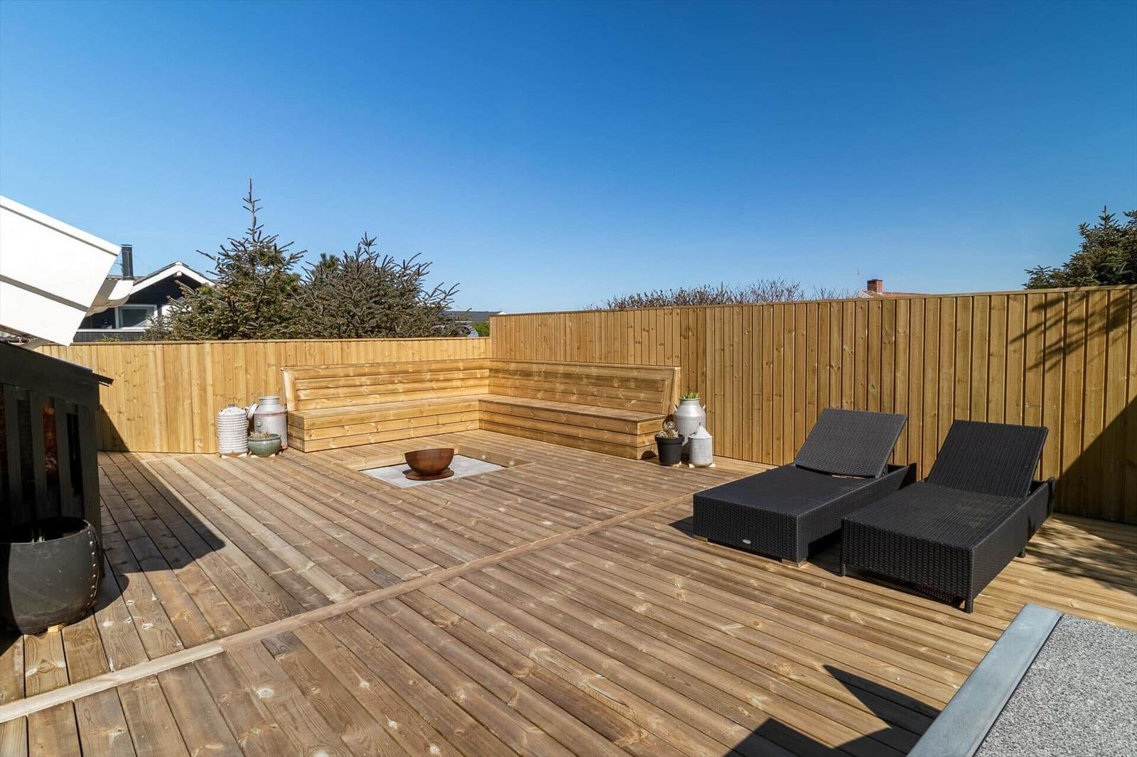 Holzterrasse mit Liegen, Sitzbank und Feuerkorb. Umzäunt mit Holz und Blick auf Bäume.