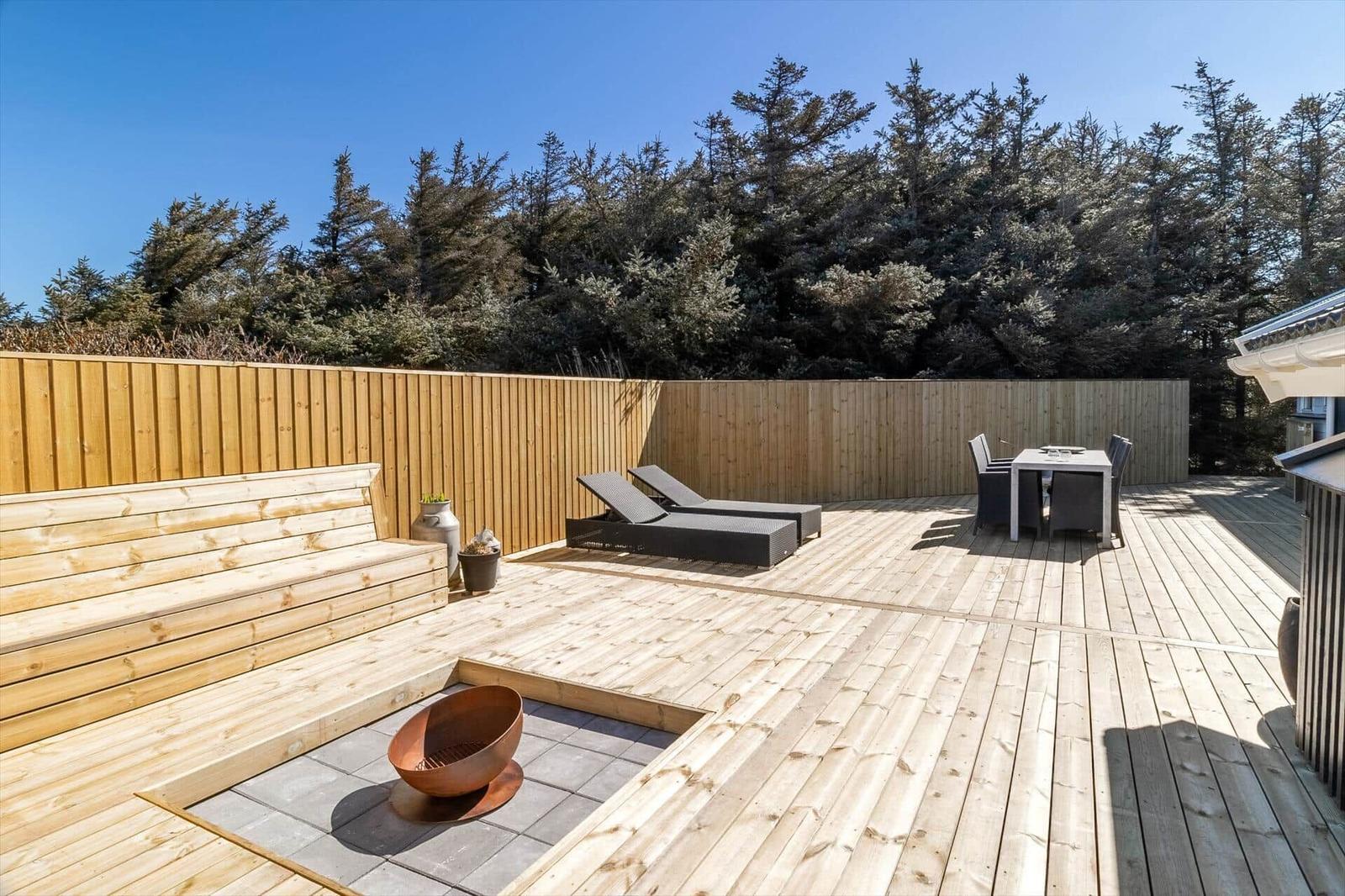 Holzterrasse mit Liegen, Esstisch und integriertem Feuerkorb. Hintergrund: Holzzaun und Nadelbäume.