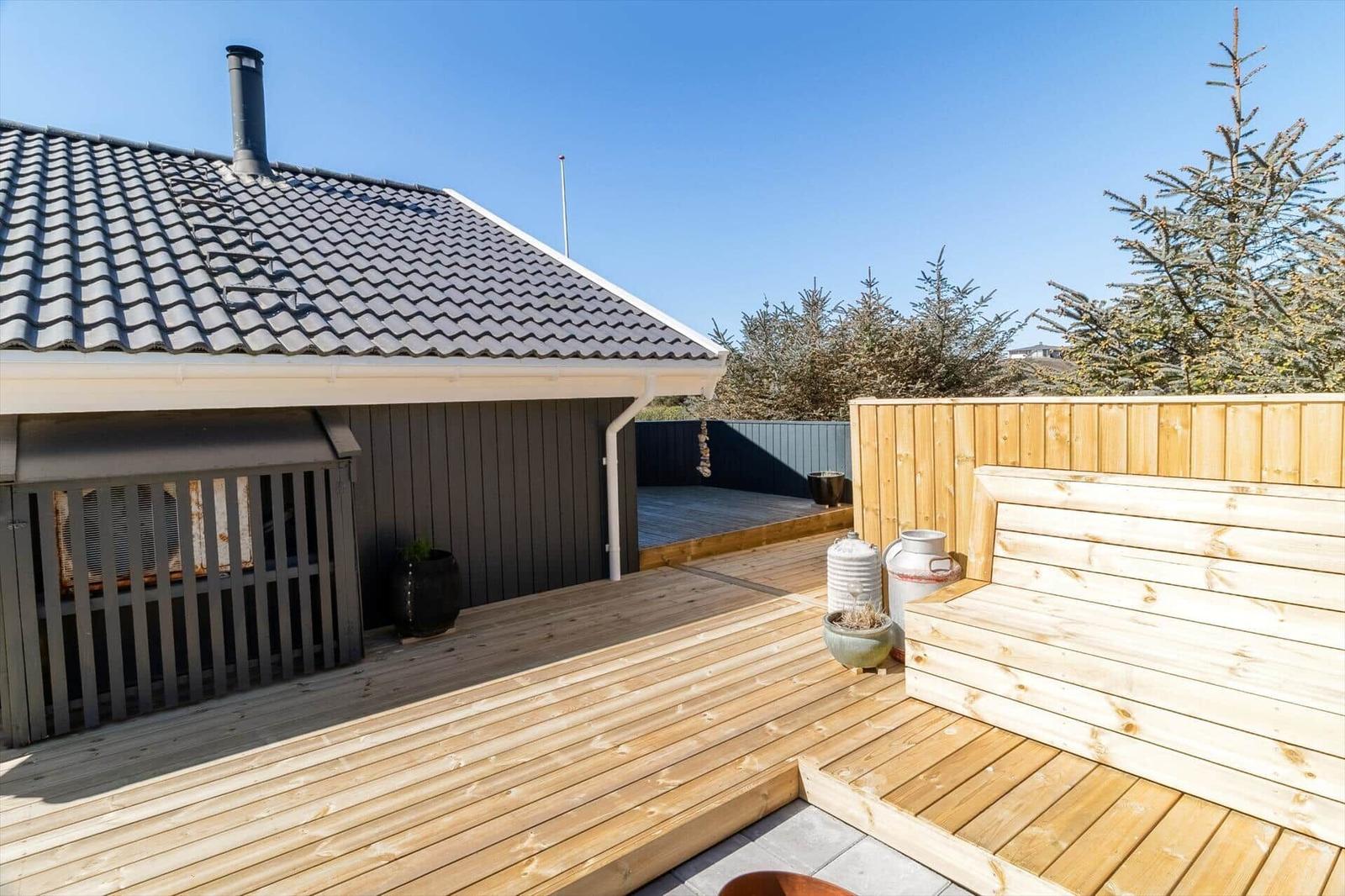 Holzterrasse mit Sitzbank, Grill und Blick auf Bäume.