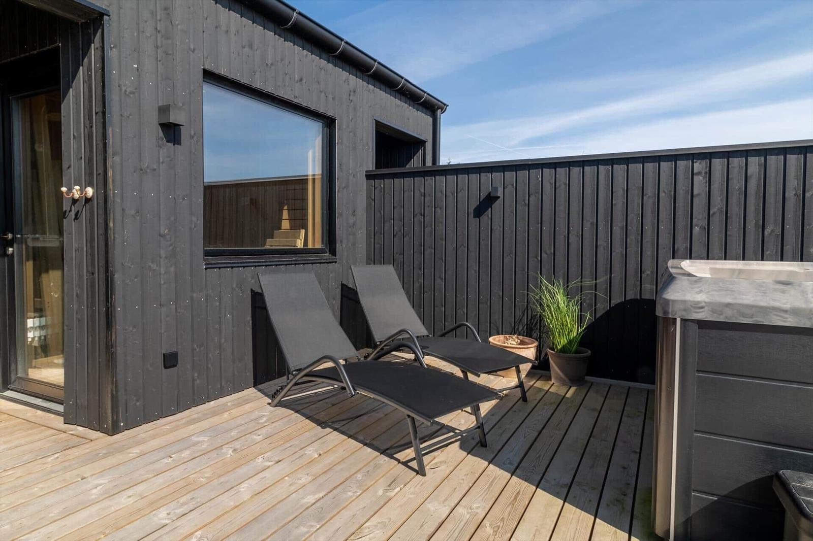 Terrasse mit zwei Liegestühlen, Holzboden und Whirlpool. Schwarze Holzwand und Fenster sichtbar.