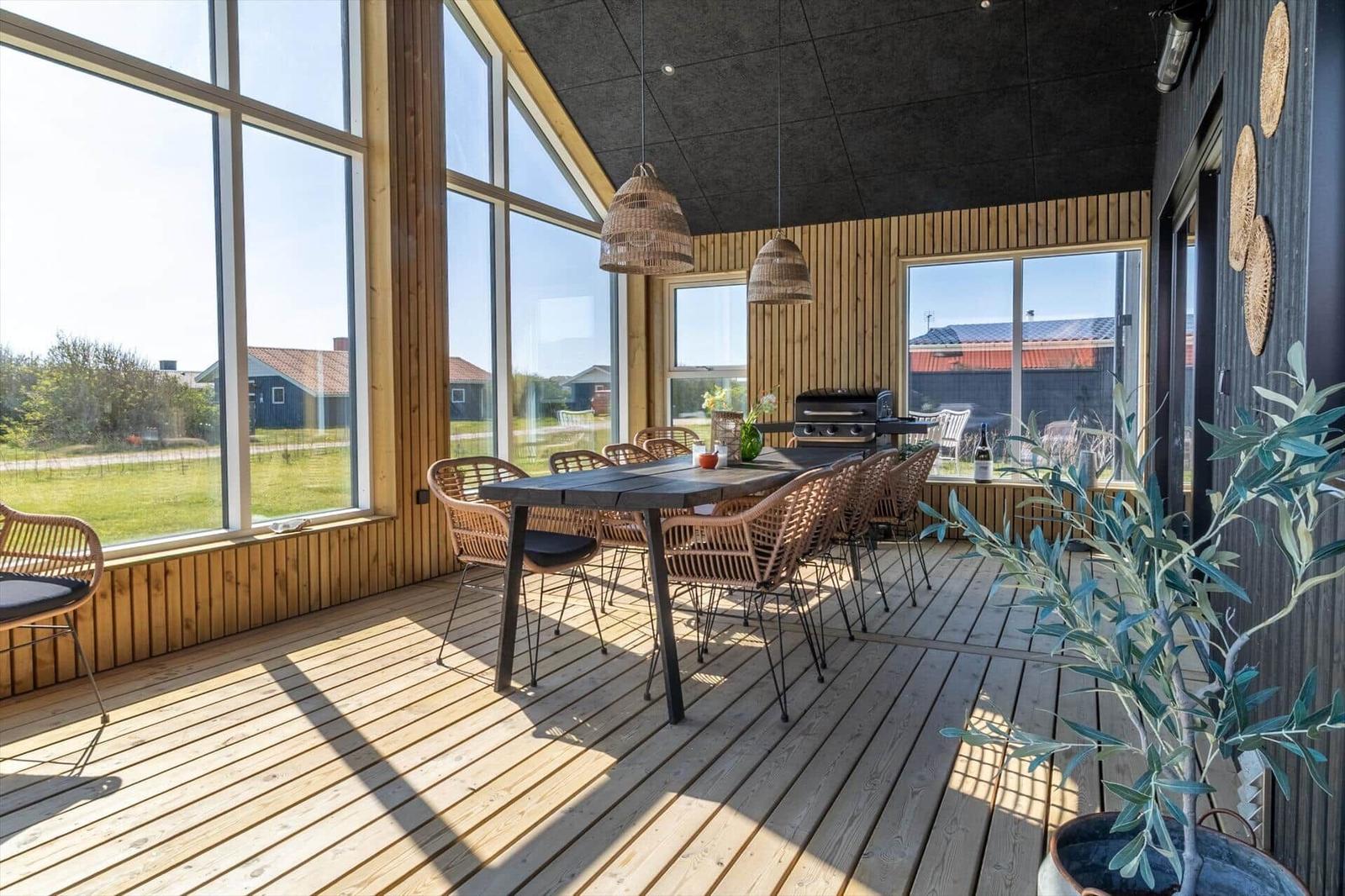 Terrasse mit Esstisch, Stühlen und Grill. Große Fenster, Holzboden und Pflanze.