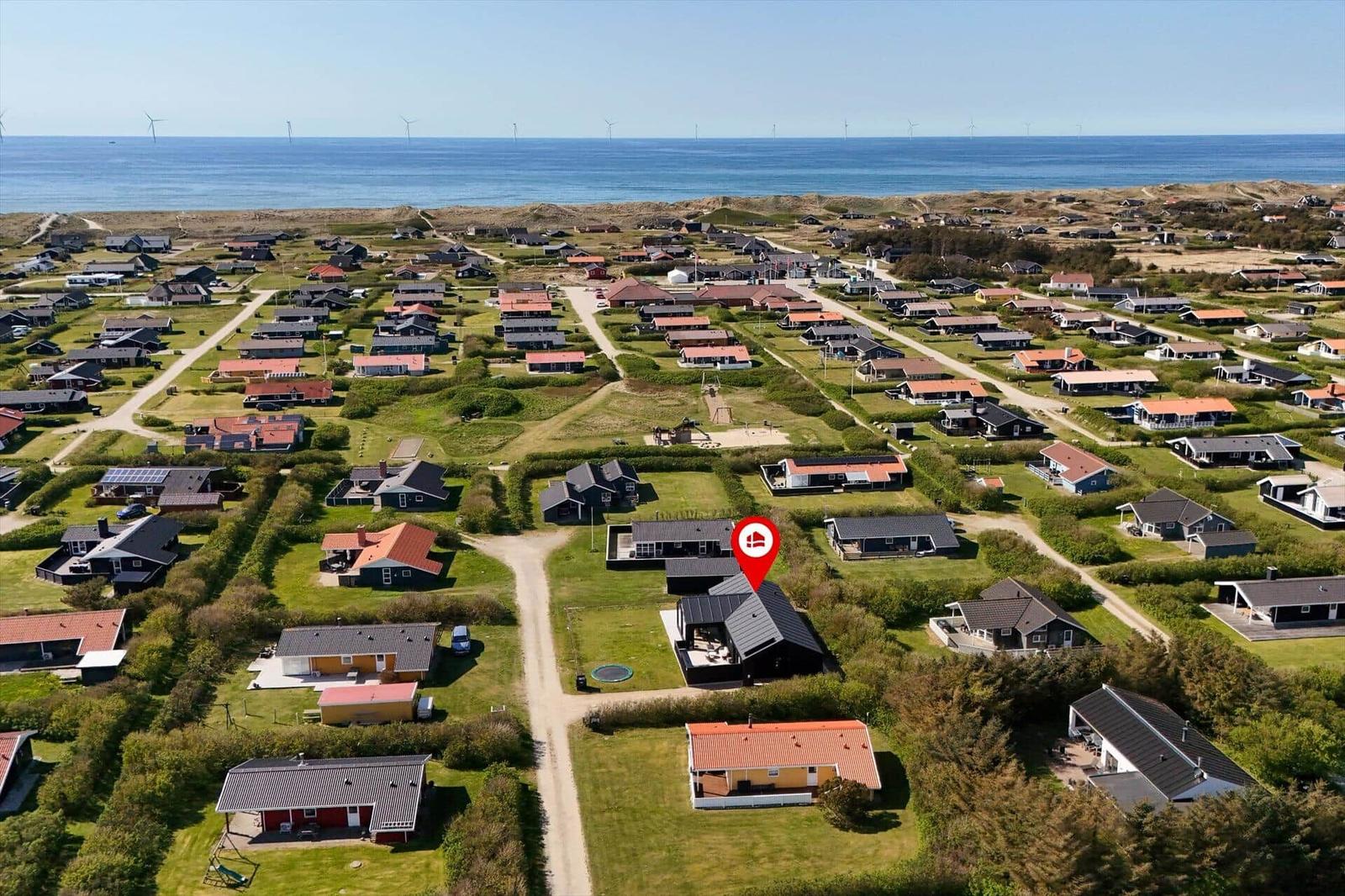 Aerial view of a coastal village with houses, greenery, and wind turbines near the sea.