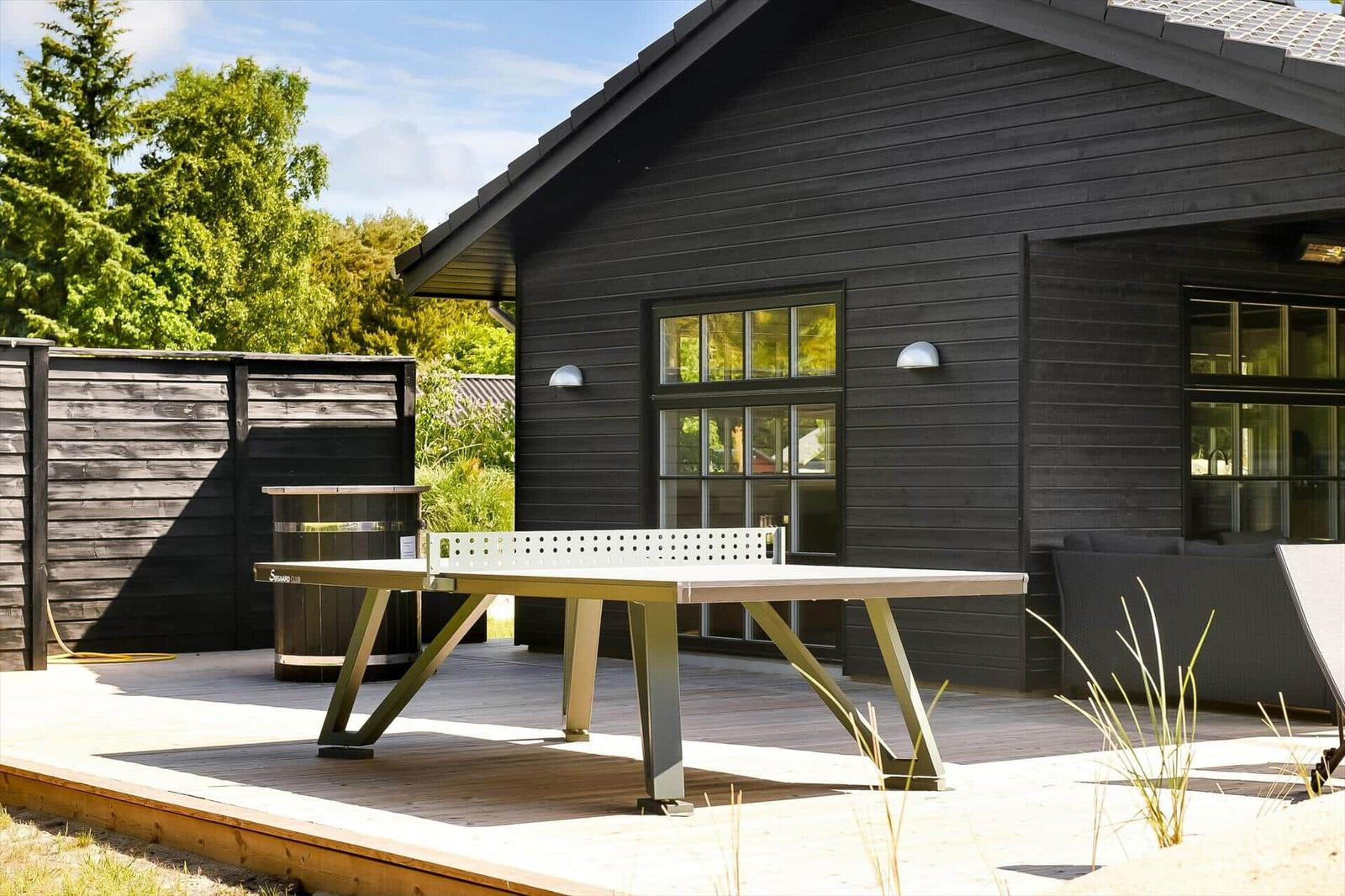 Pingpong-Tisch auf Terrasse vor schwarzem Holzhaus mit Garten und Fenstern.