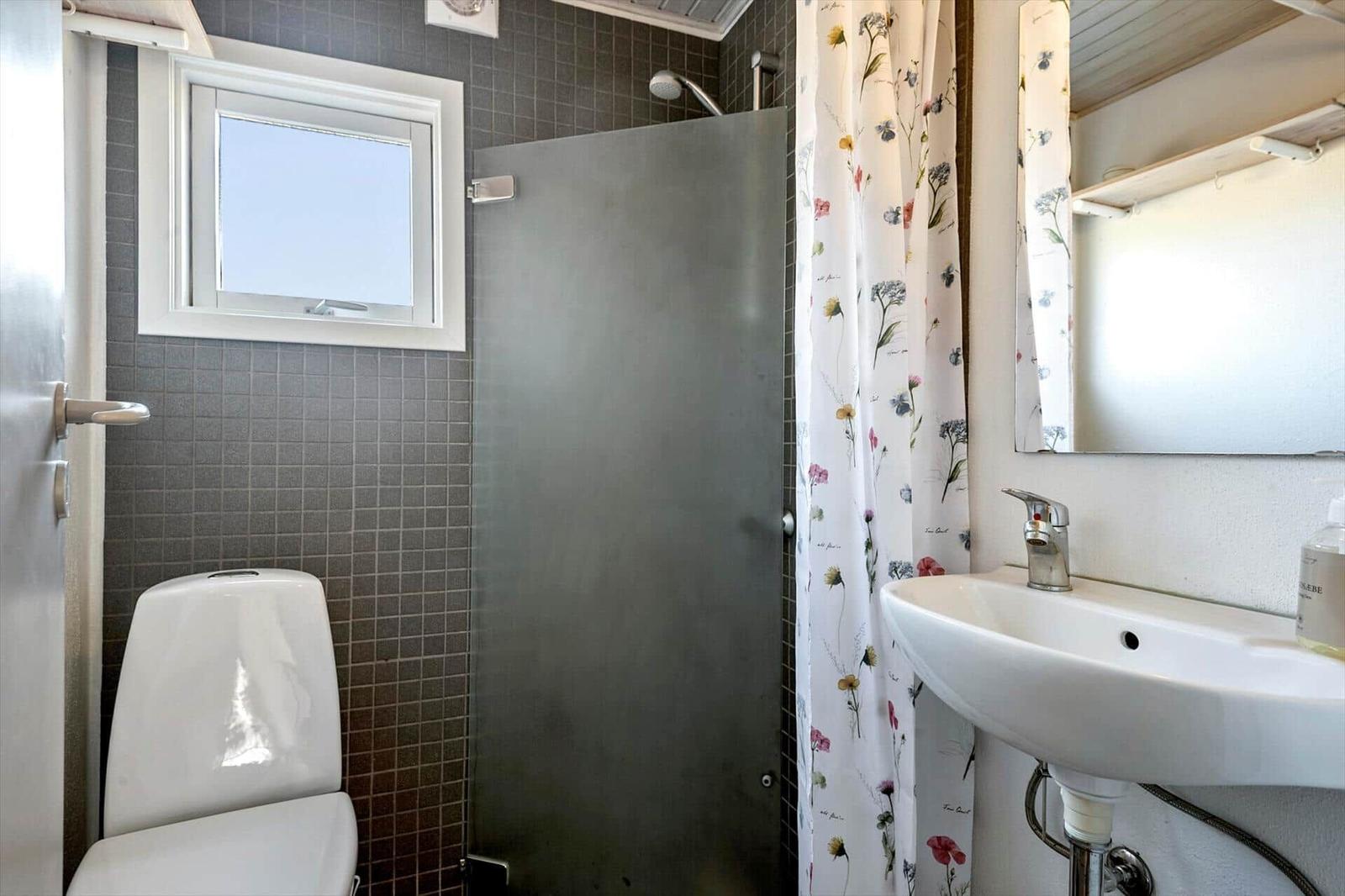 Badezimmer mit Dusche, Toilette und Waschbecken. Fenster und Blumenvorhang sichtbar.