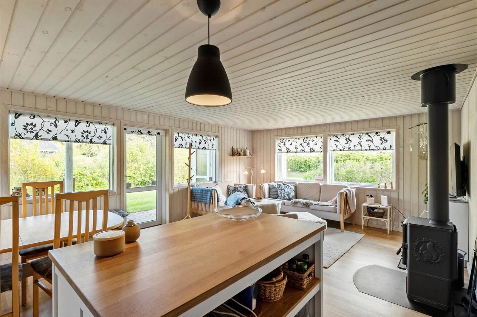 Living and dining area with wooden table, sofa, and wood stove. Windows with floral blinds.