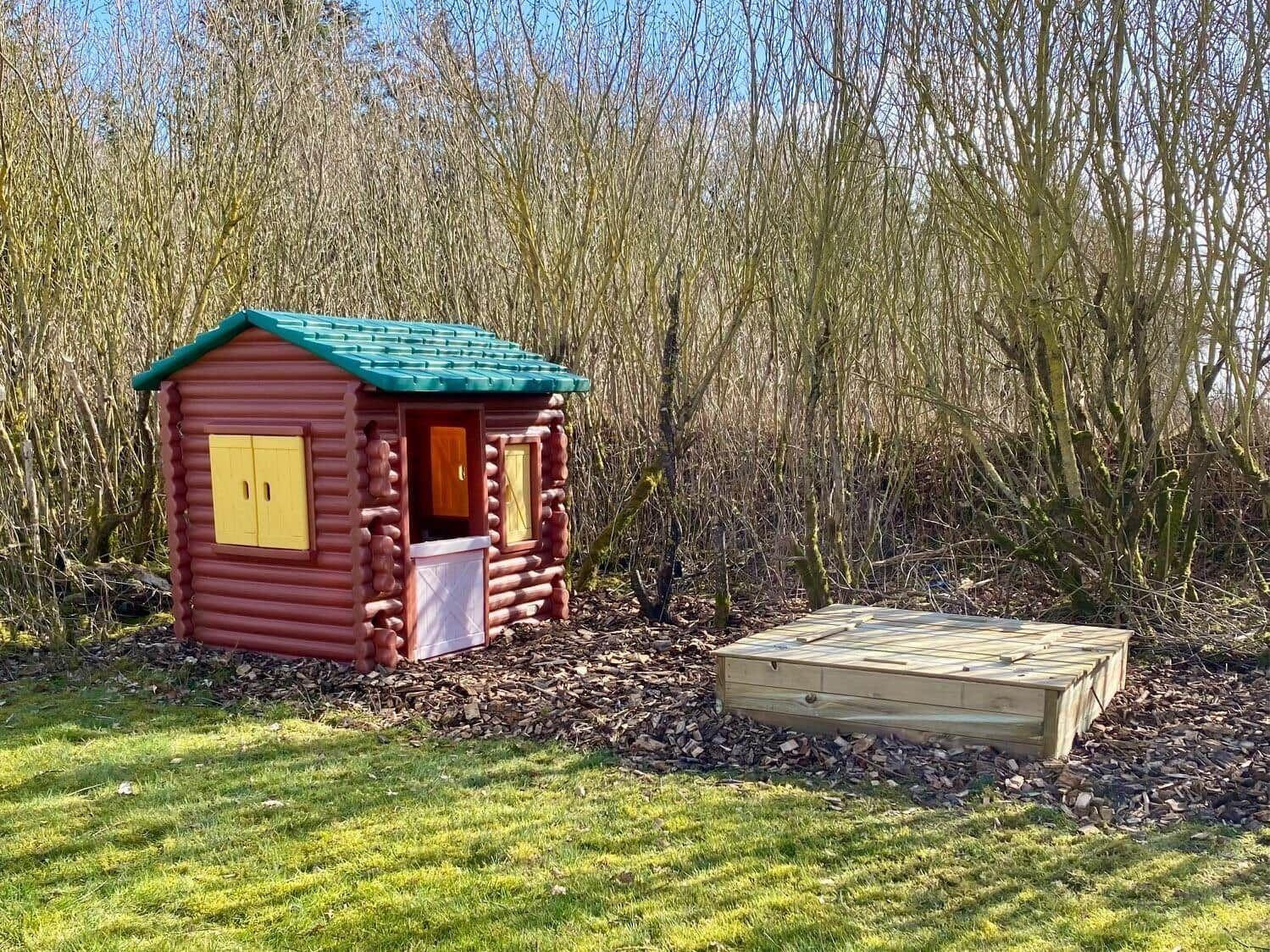 Playhouse with green roof and yellow windows next to wooden platform in the woods.