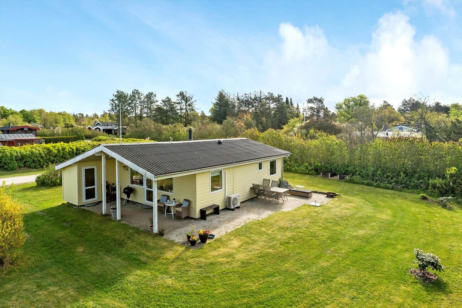 A yellow house with terrace, grill, and garden. Surrounded by trees and grass.