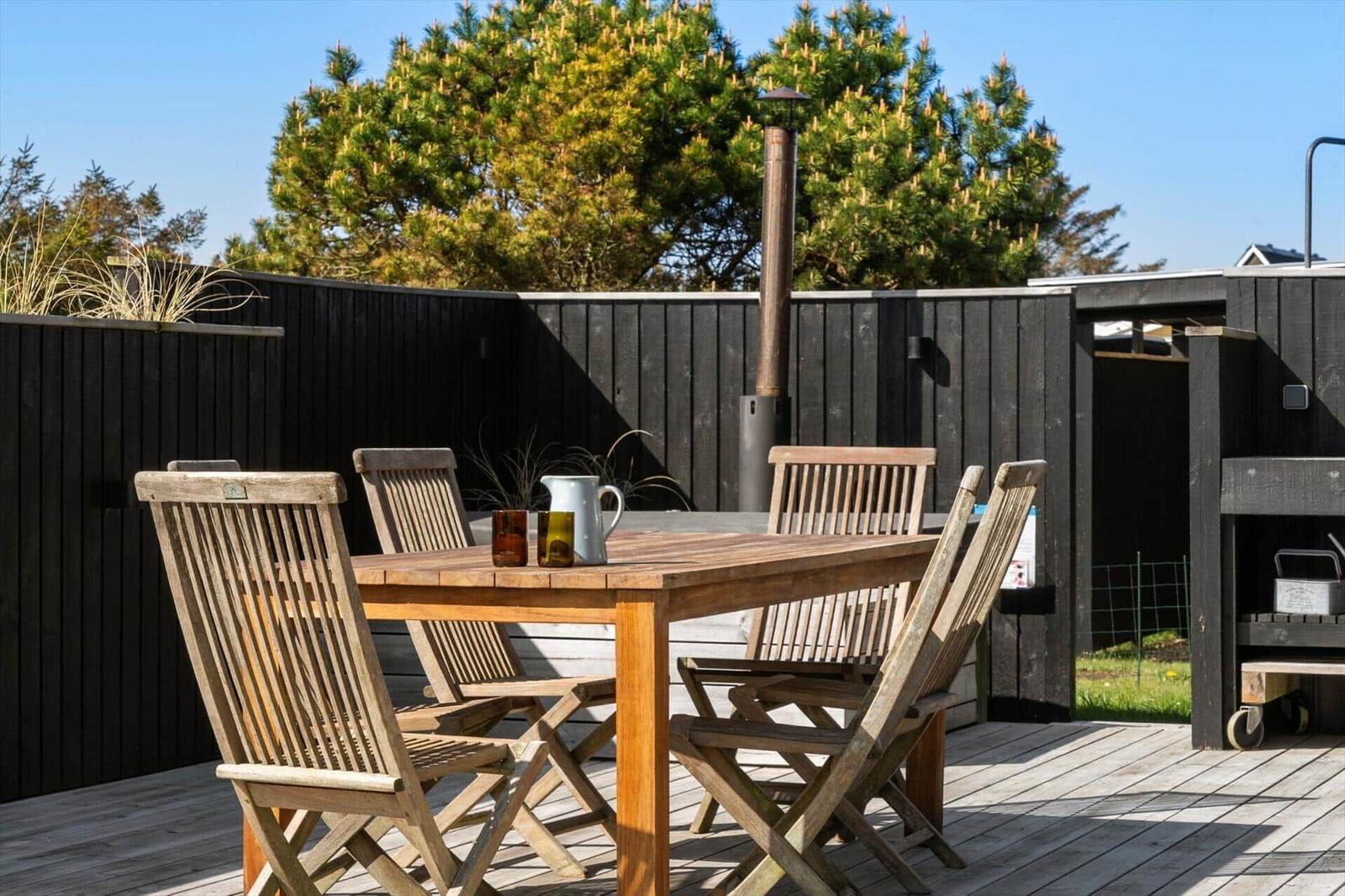 Holztisch mit Stühlen auf Terrasse, Grill und Hinterhof mit Bäumen.