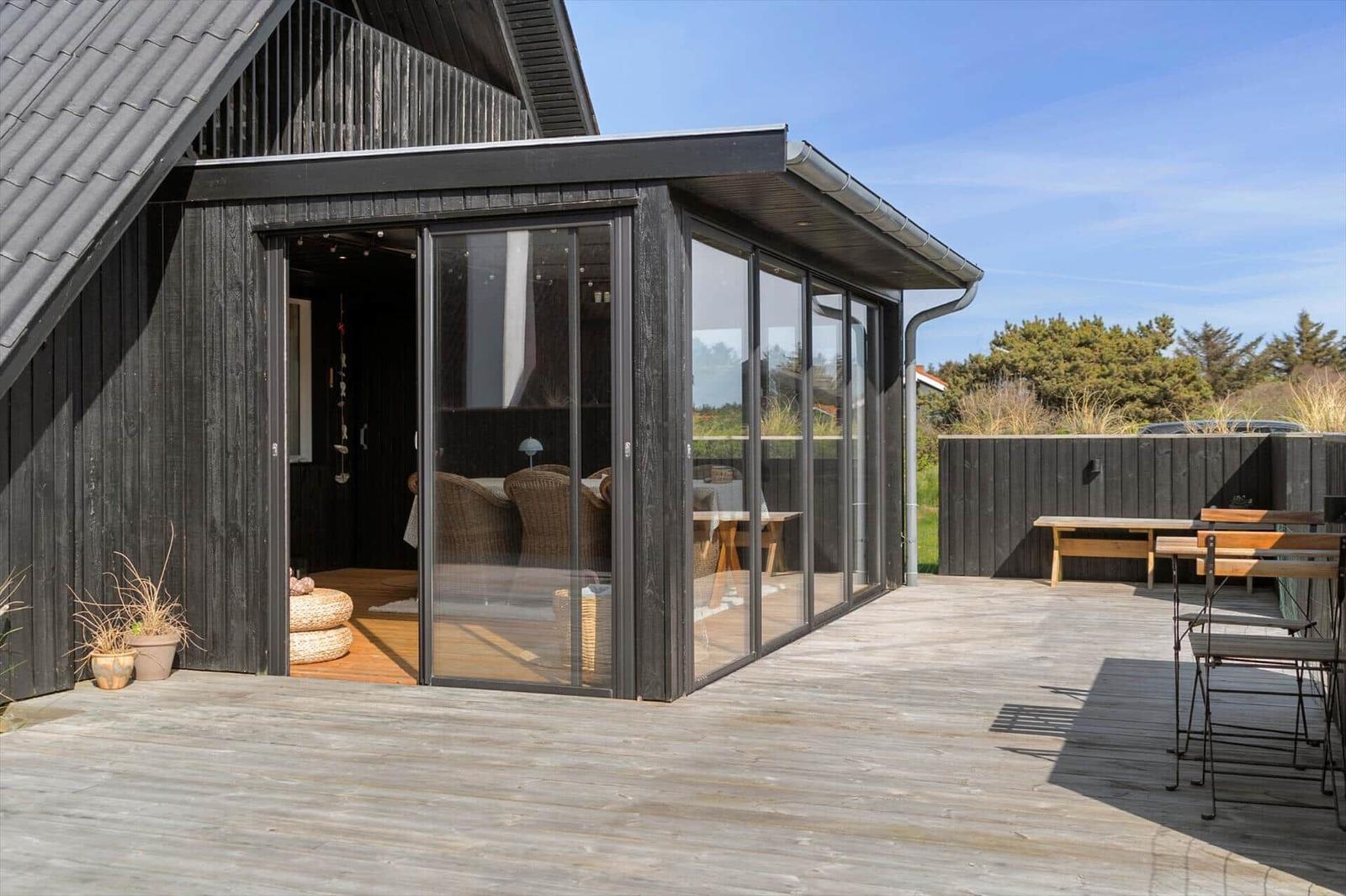 Terrasse mit Glaswand und Sitzgelegenheiten vor einem Haus mit dunklem Holzverkleidung.