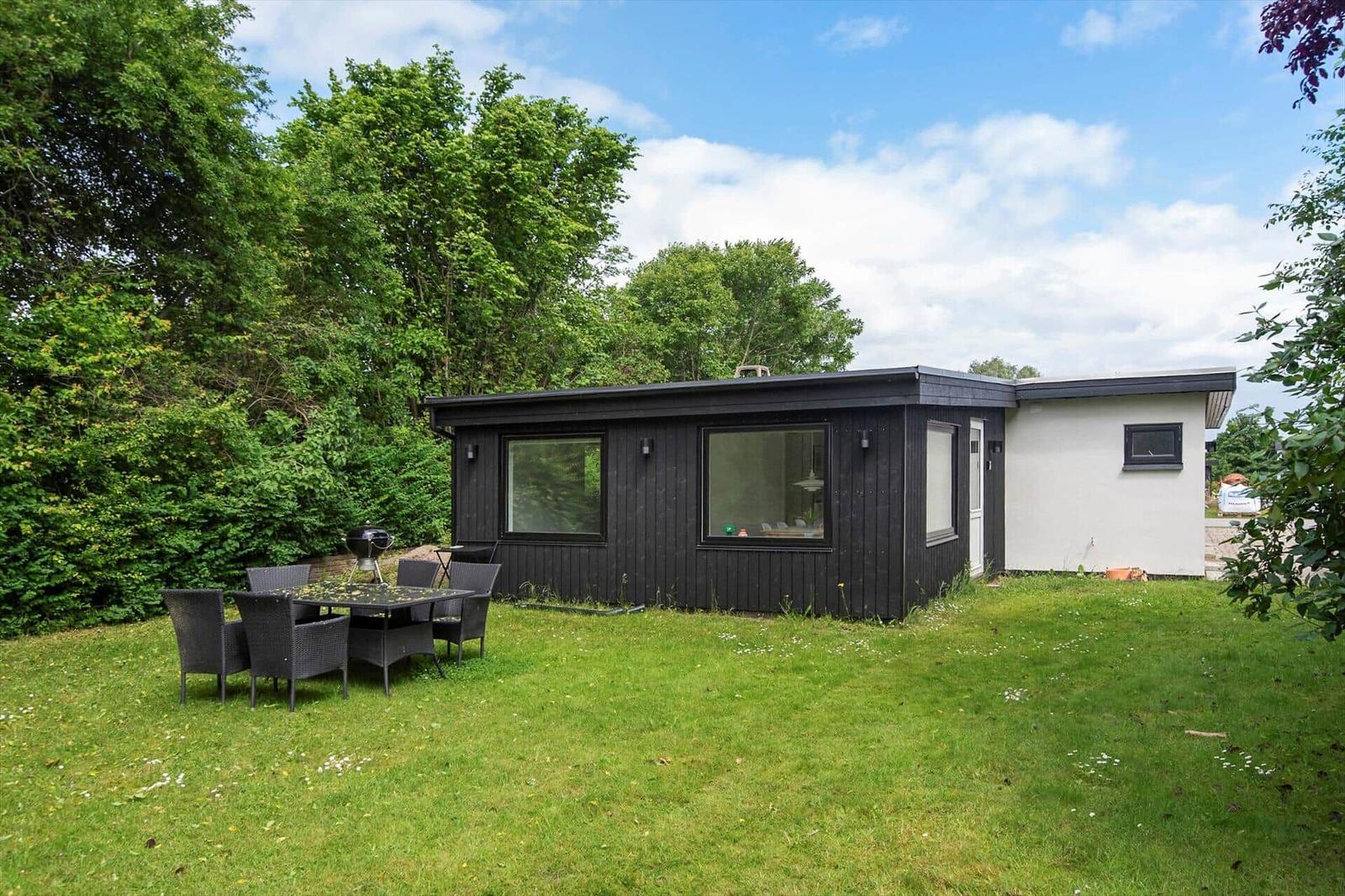 Ein Haus mit dunkler Fassade und weißem Anbau. Auf dem Rasen steht ein Grill und ein Tisch mit Stühlen.