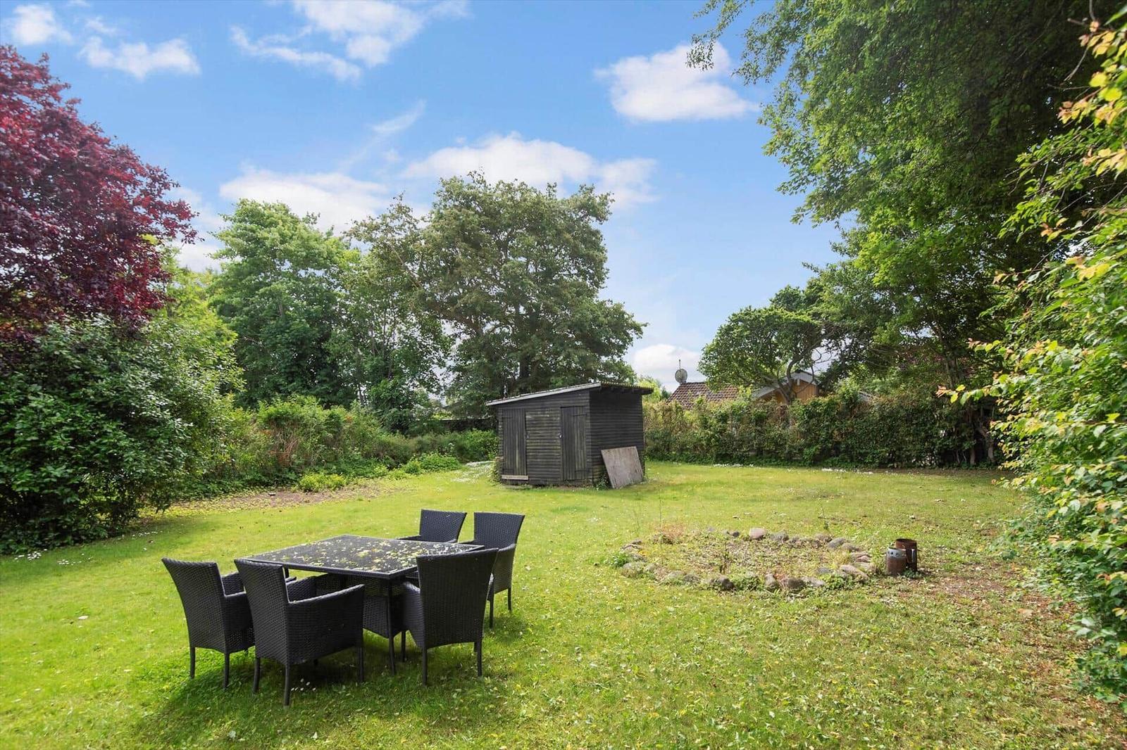 Grüner Garten mit Tisch und Stühlen, kleiner Holzschuppen und Bäumen im Hintergrund.