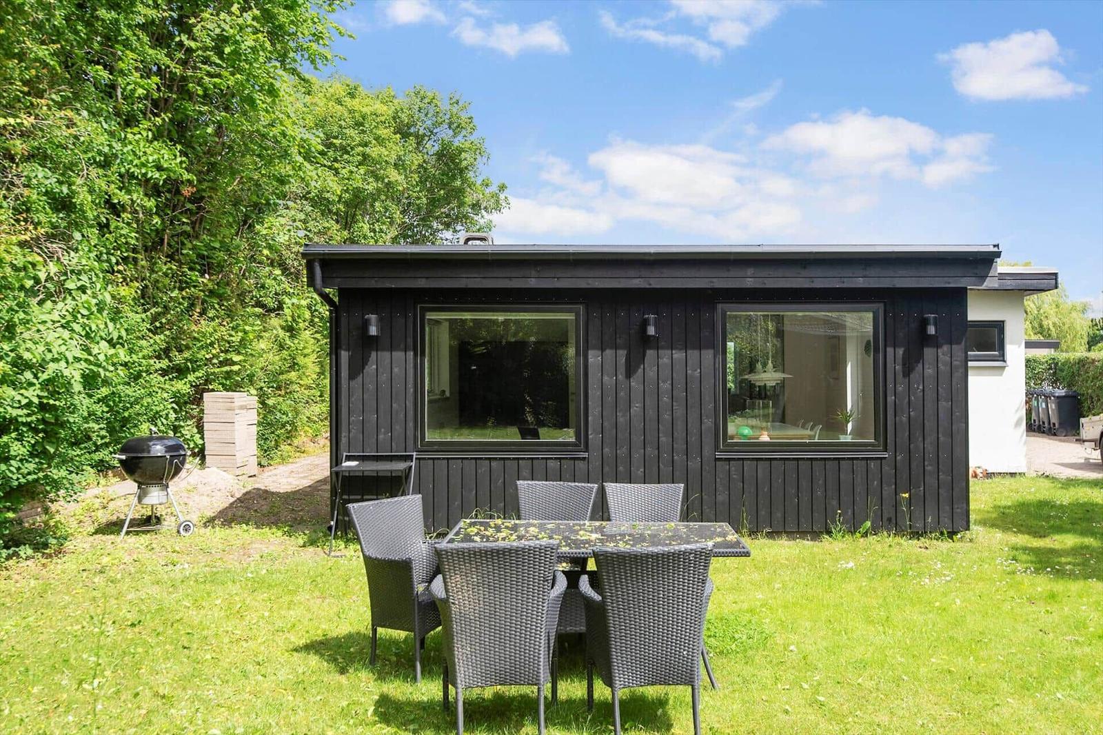 Schwarzes Holzhaus mit Gartenmöbeln und Grill. Grün umgeben unter blauem Himmel.