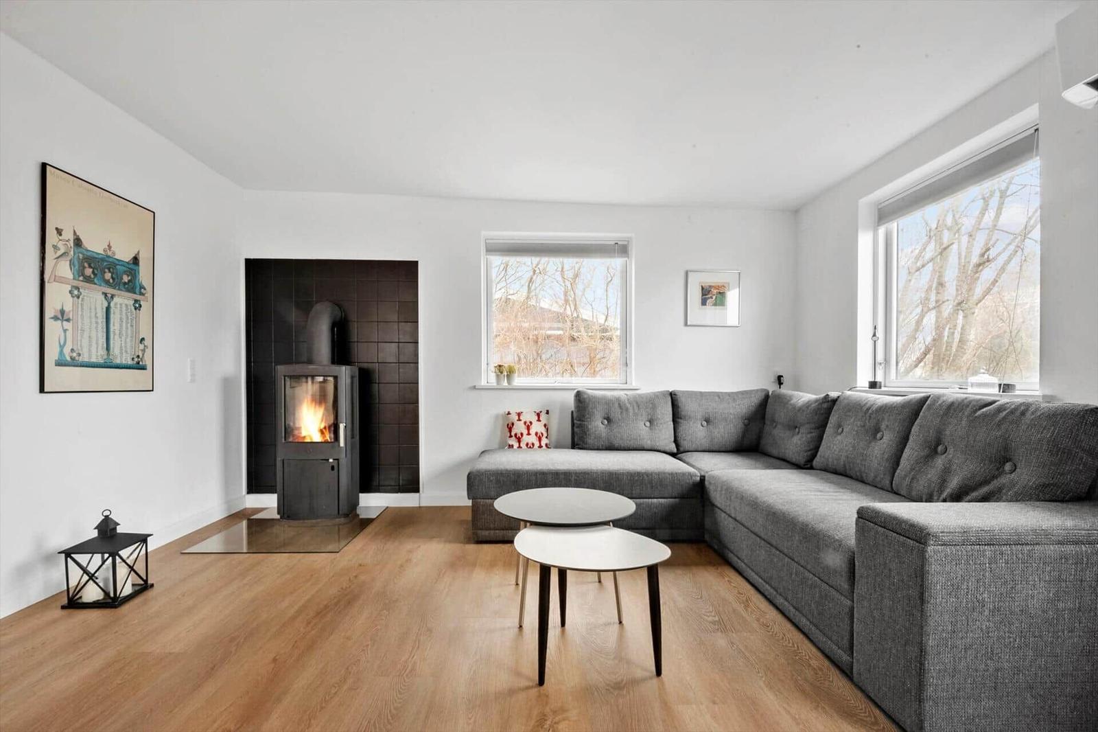 Wohnzimmer mit grauem Sofa, Kamin und Holzboden. Fenster mit Blick auf Bäume.