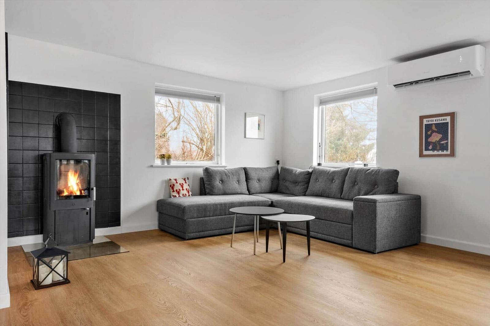 Wohnzimmer mit grauem Sofa, Kamin und Holzboden. Zwei Fenster und Wandklimaanlage.