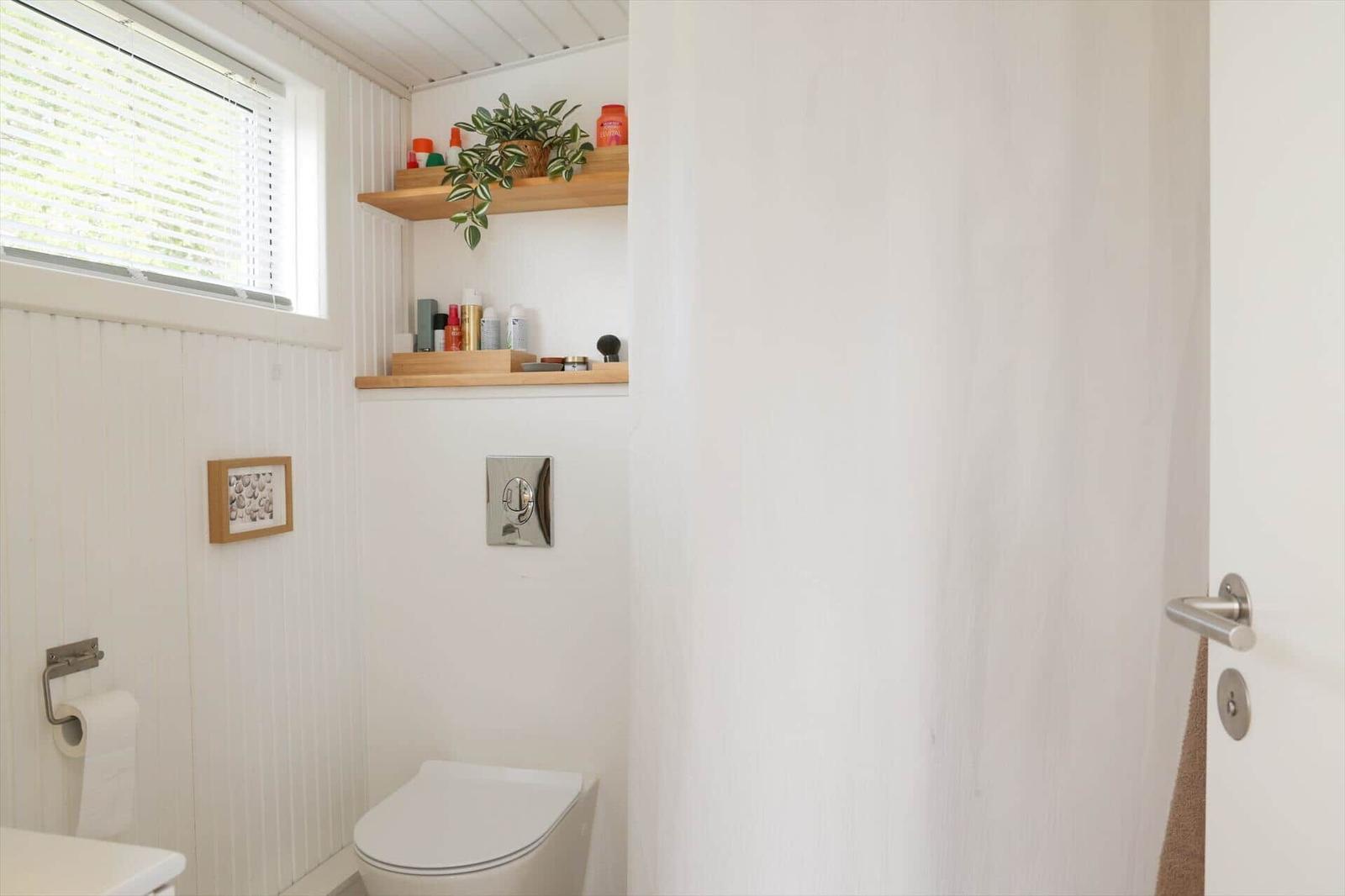 Badezimmer mit Toilette, Fenster und Holzregal mit Pflanze und Pflegeprodukten.