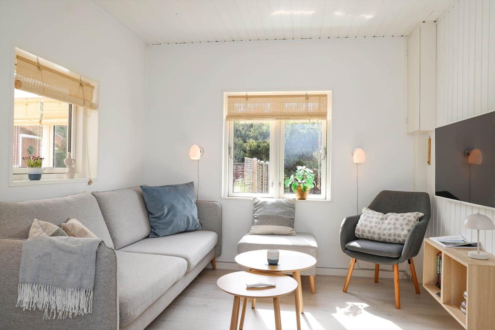 Wohnzimmer mit Sofa, Sessel und zwei Tischen. Fenster mit Jalousien und Pflanzen.