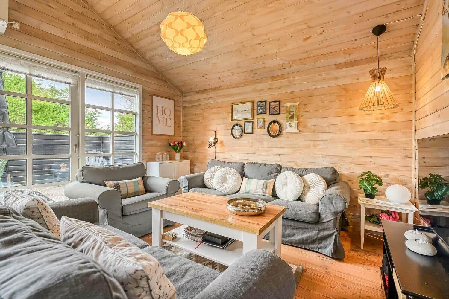 Wohnzimmer mit Holzwänden, grauen Sofas und Holztisch. Fenster mit Blick auf Balkon.