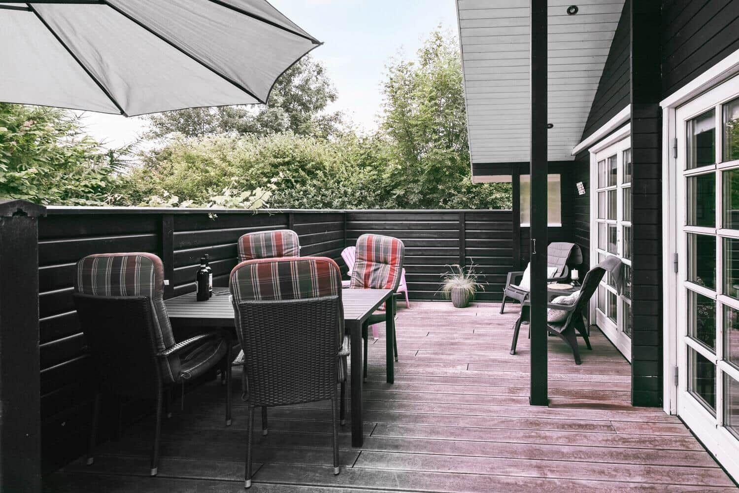 Terrasse mit Tisch, Stühlen und Sonnenschirm. Holzdecke und Pflanzen im Hintergrund.