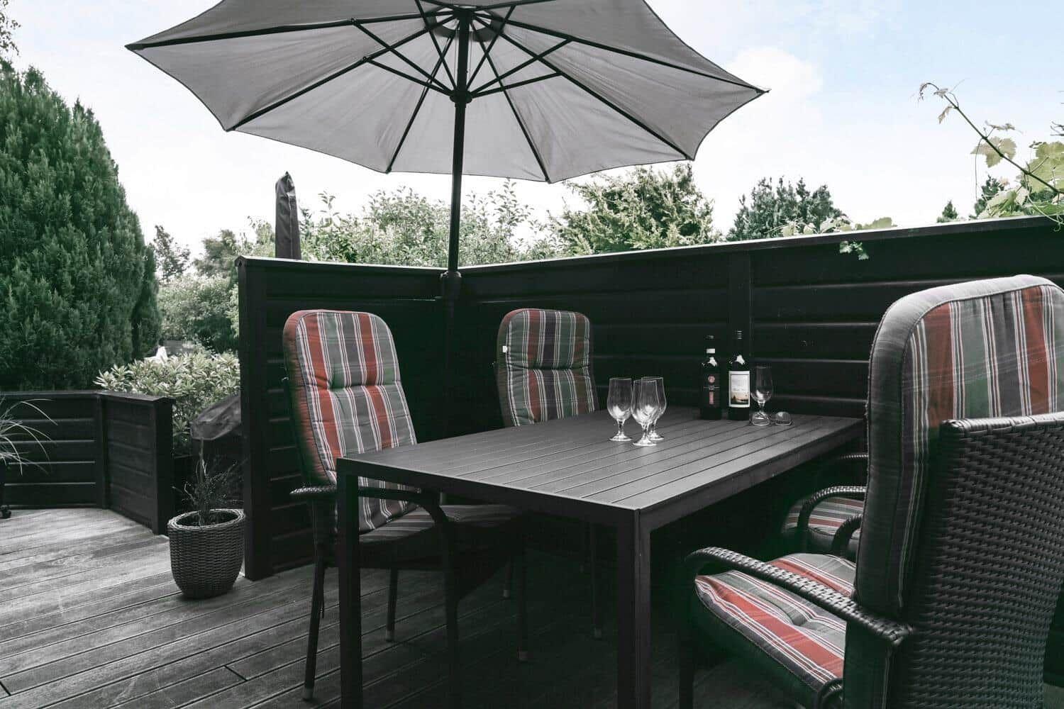Terrasse mit Tisch, Stühlen und Sonnenschirm. Hintergrund: dunkle Holzumrandung und Bäume.