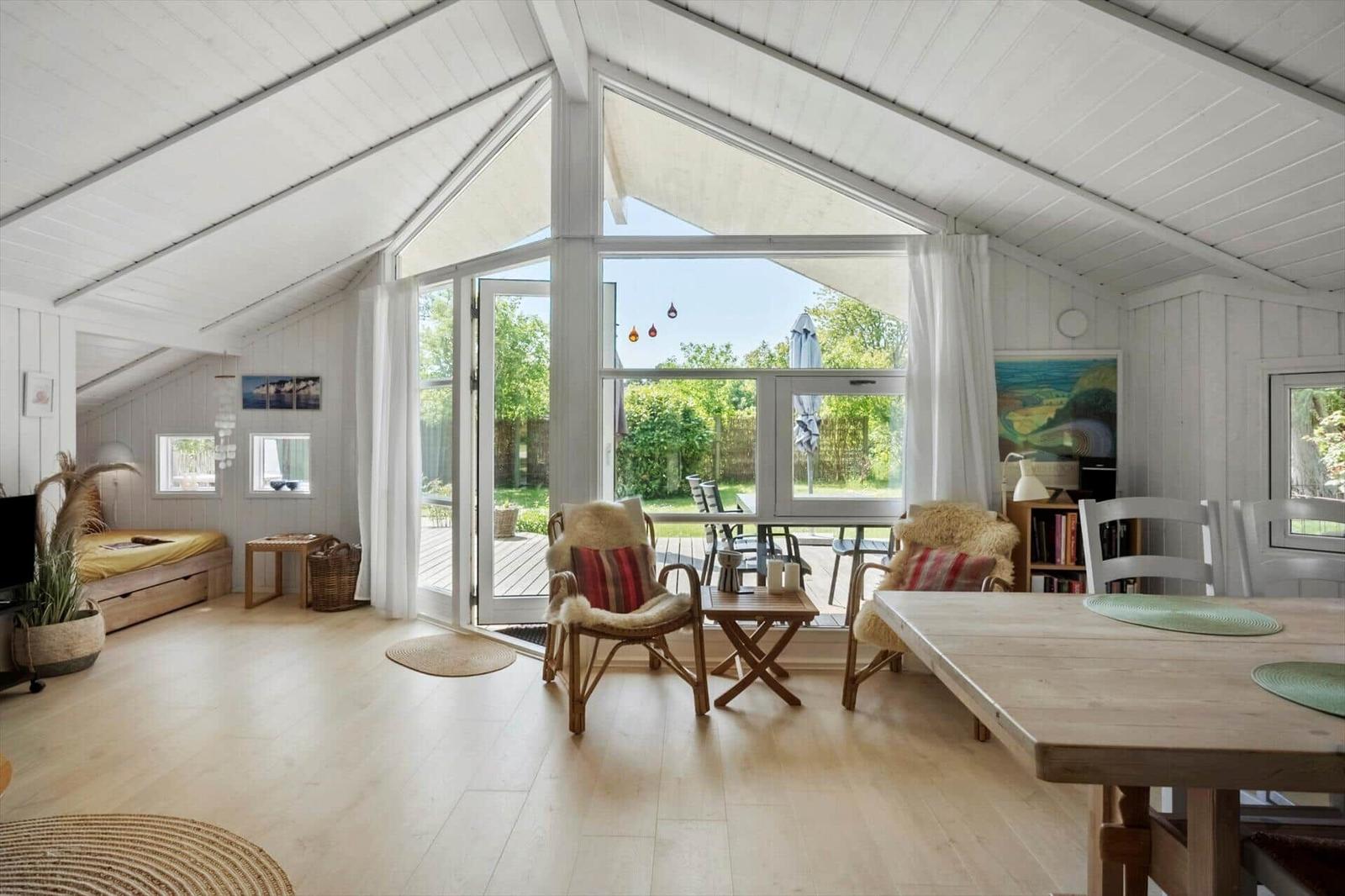 Ein Raum mit hölzernem Boden, großem Fenster und Sitzgelegenheiten. Draußen ist eine Terrasse mit Garten.