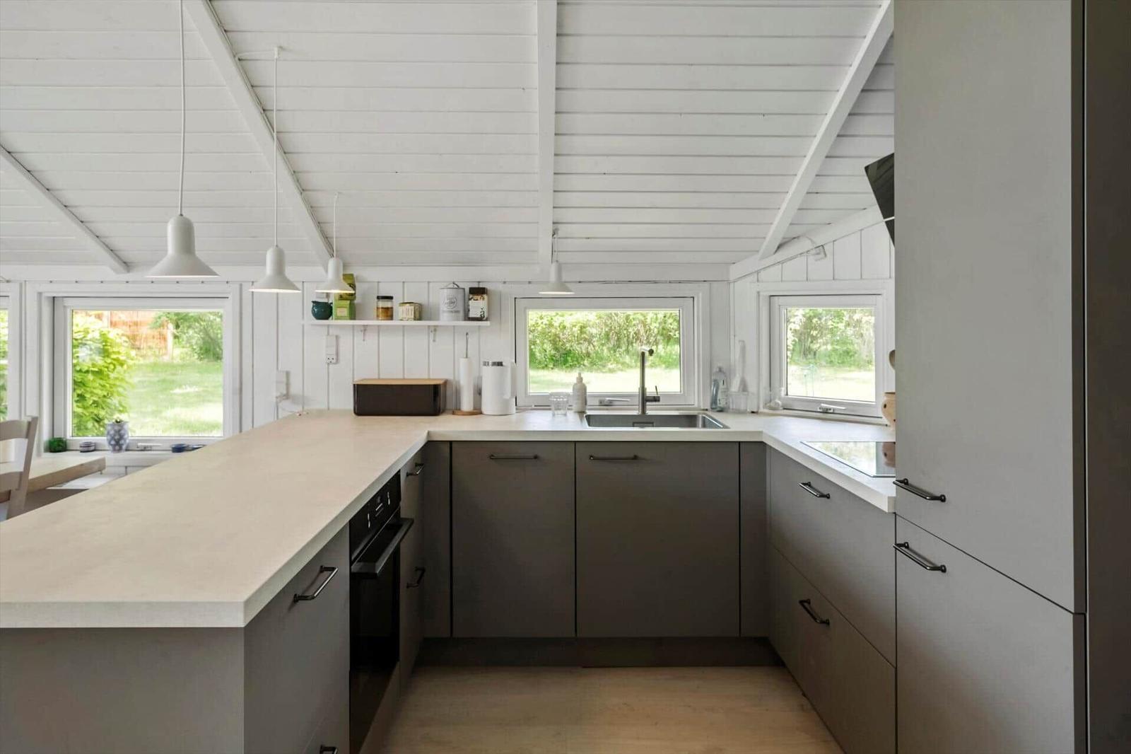 Küche mit Arbeitsplatte, Spüle und Fenstern. Weiße Decke und graue Schränke.