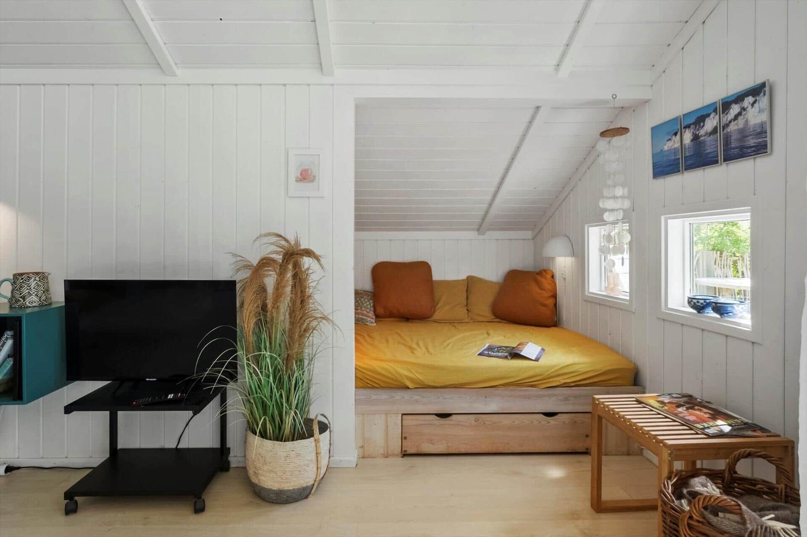 Schlafbereich mit Bett, Fenster und Wandbildern. Fernseher und Pflanze im Vordergrund.