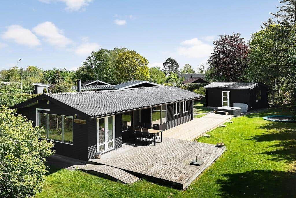 Black vacation house with terrace, garden, and pool. Surrounded by trees and greenery.