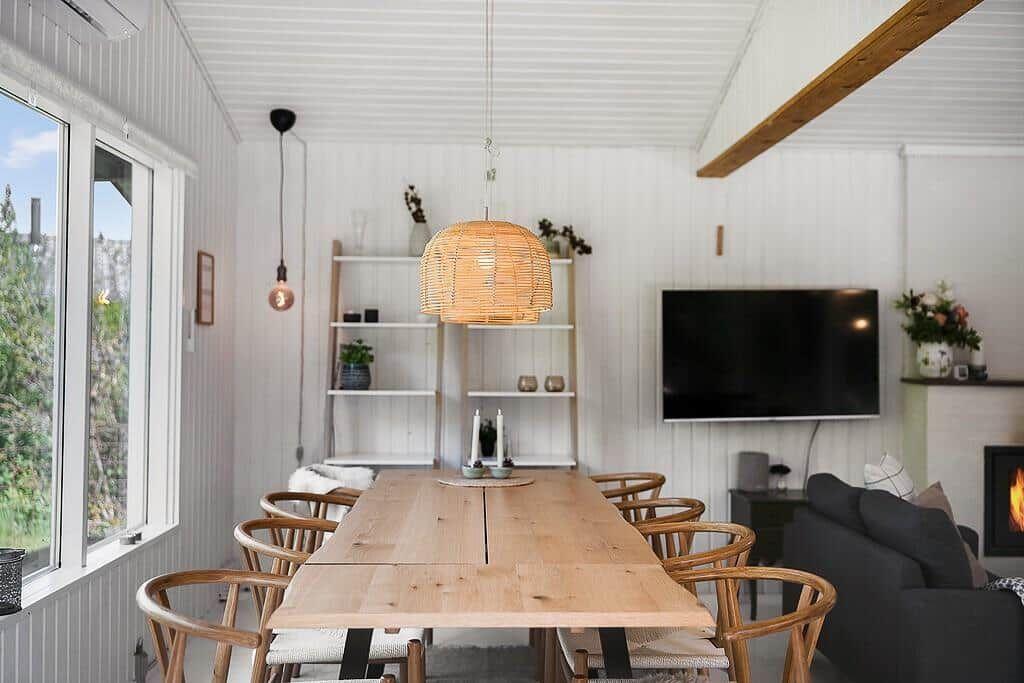 Dining room with wooden table, chairs, and TV. Large windows, fireplace, and ceiling beam.