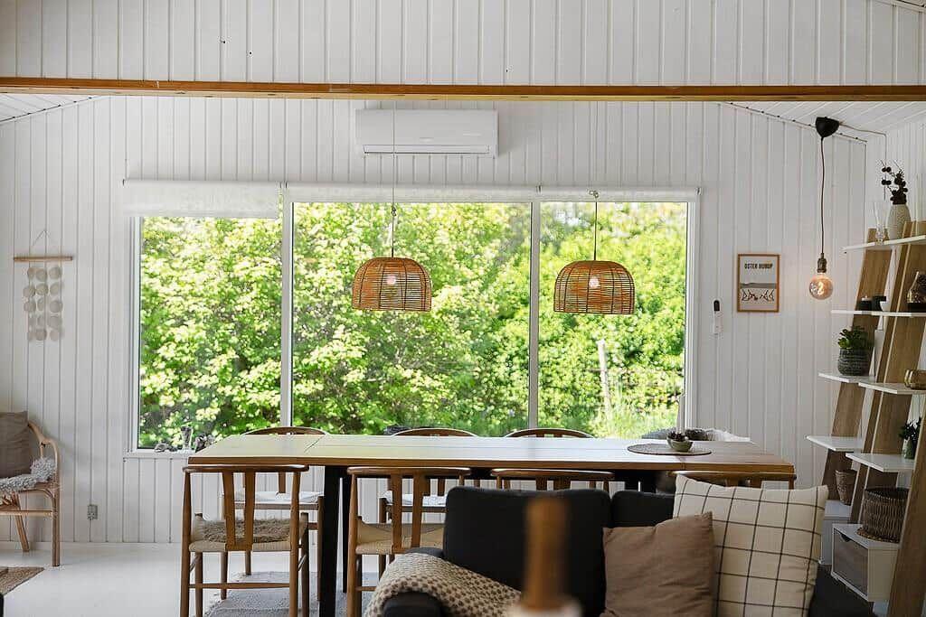 Dining room with wooden table, chairs, and view of green trees through large windows.