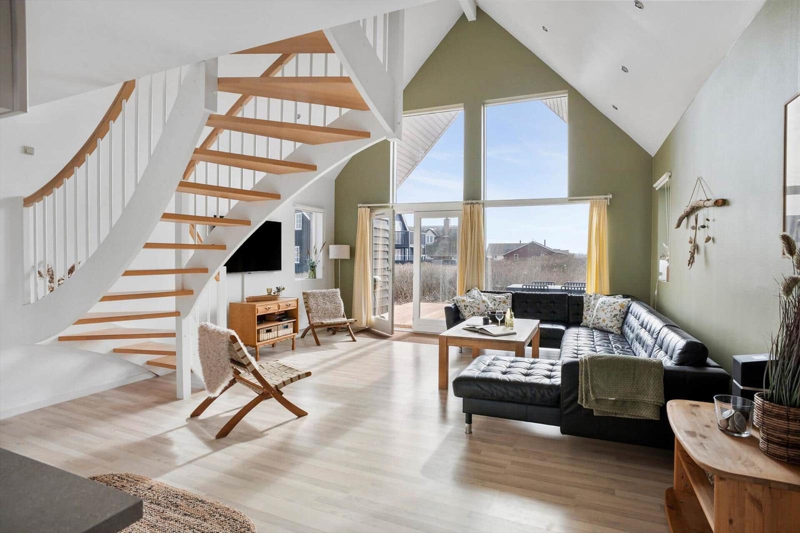 Wohnzimmer mit schwarzem Sofa, Holztreppe und großen Fenstern zur Aussicht.