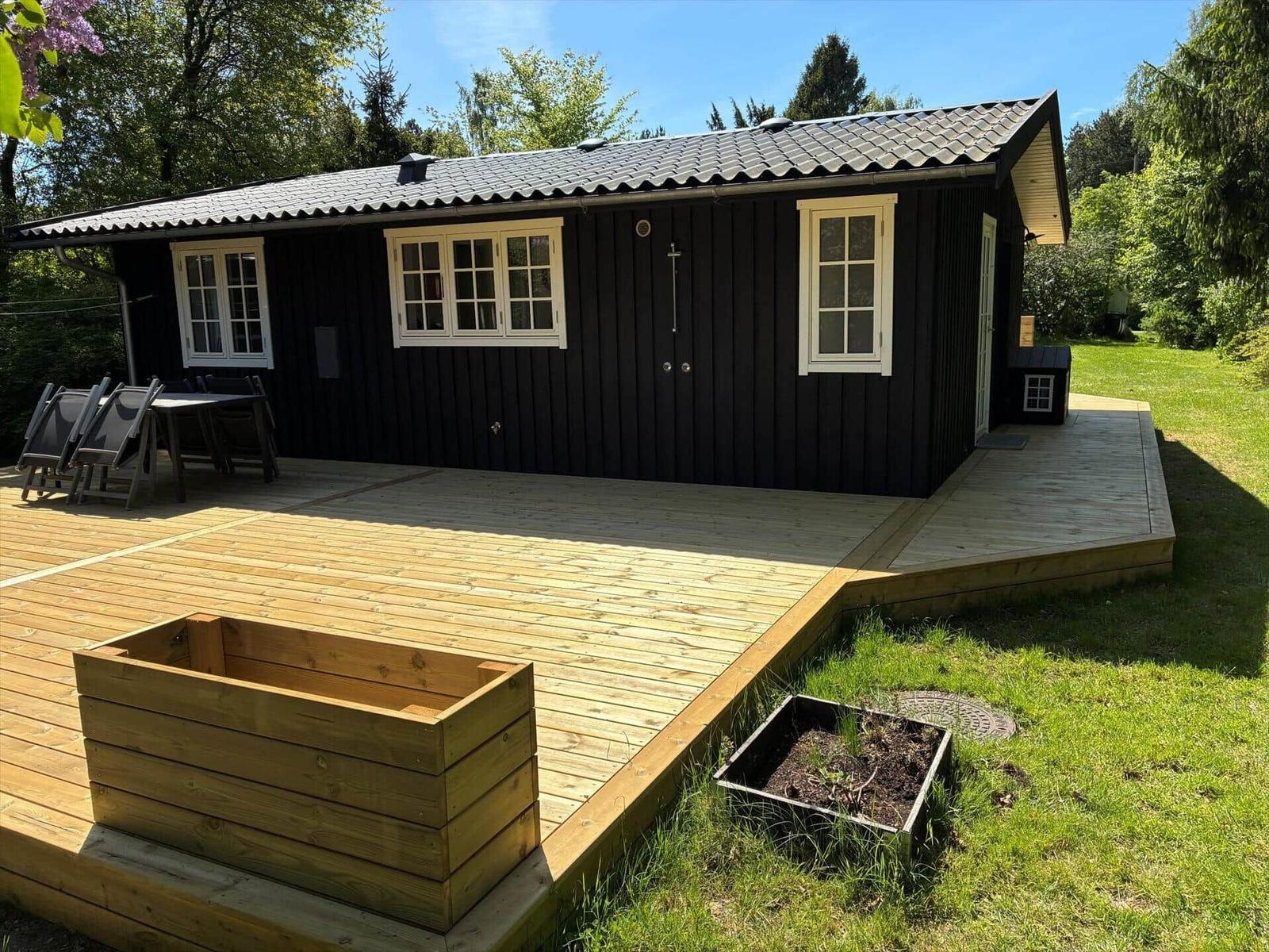 Black cabin with deck, garden, and wooden planters. Table and chairs on the deck.