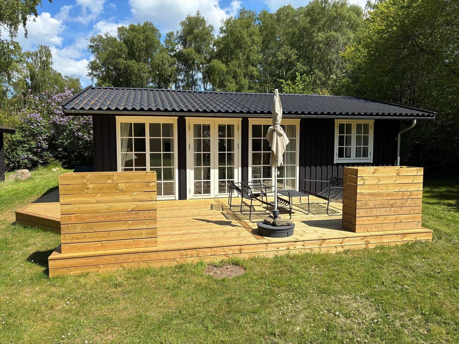 A house with wooden terrace, garden table and chairs. Background: trees and grass.
