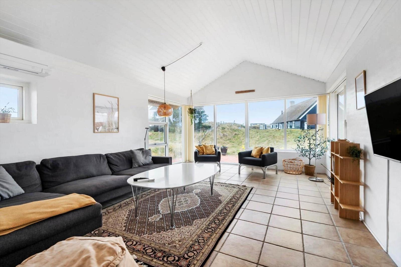 Wohnzimmer mit Sofa, Tisch und Sesseln. Große Fensterfront mit Blick auf Landschaft.