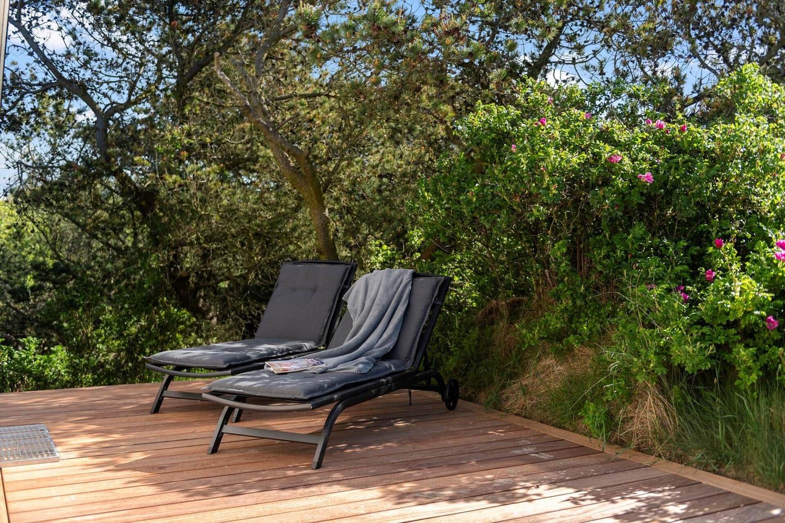 Two lounge chairs on a wooden terrace surrounded by trees and bushes with pink flowers.