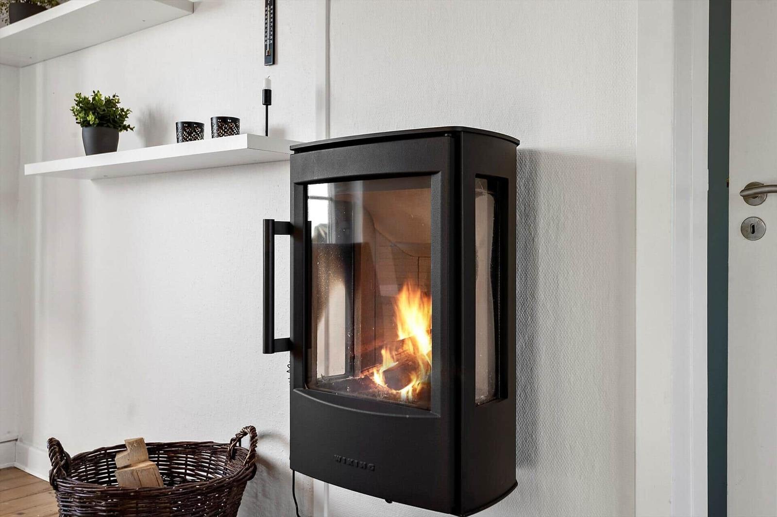 Wall-mounted stove with burning wood, beside basket with firewood and shelf with plant.