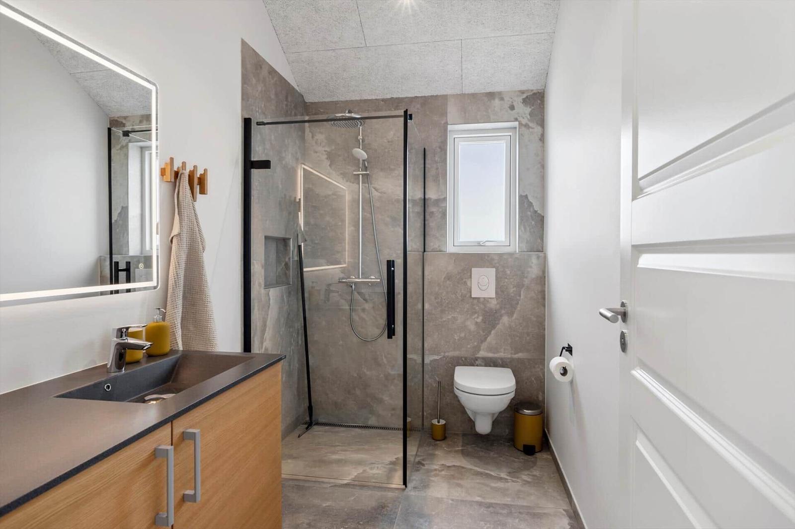 Badezimmer mit Dusche, Waschbecken und WC. Graue Fliesen, Spiegel mit Beleuchtung.
