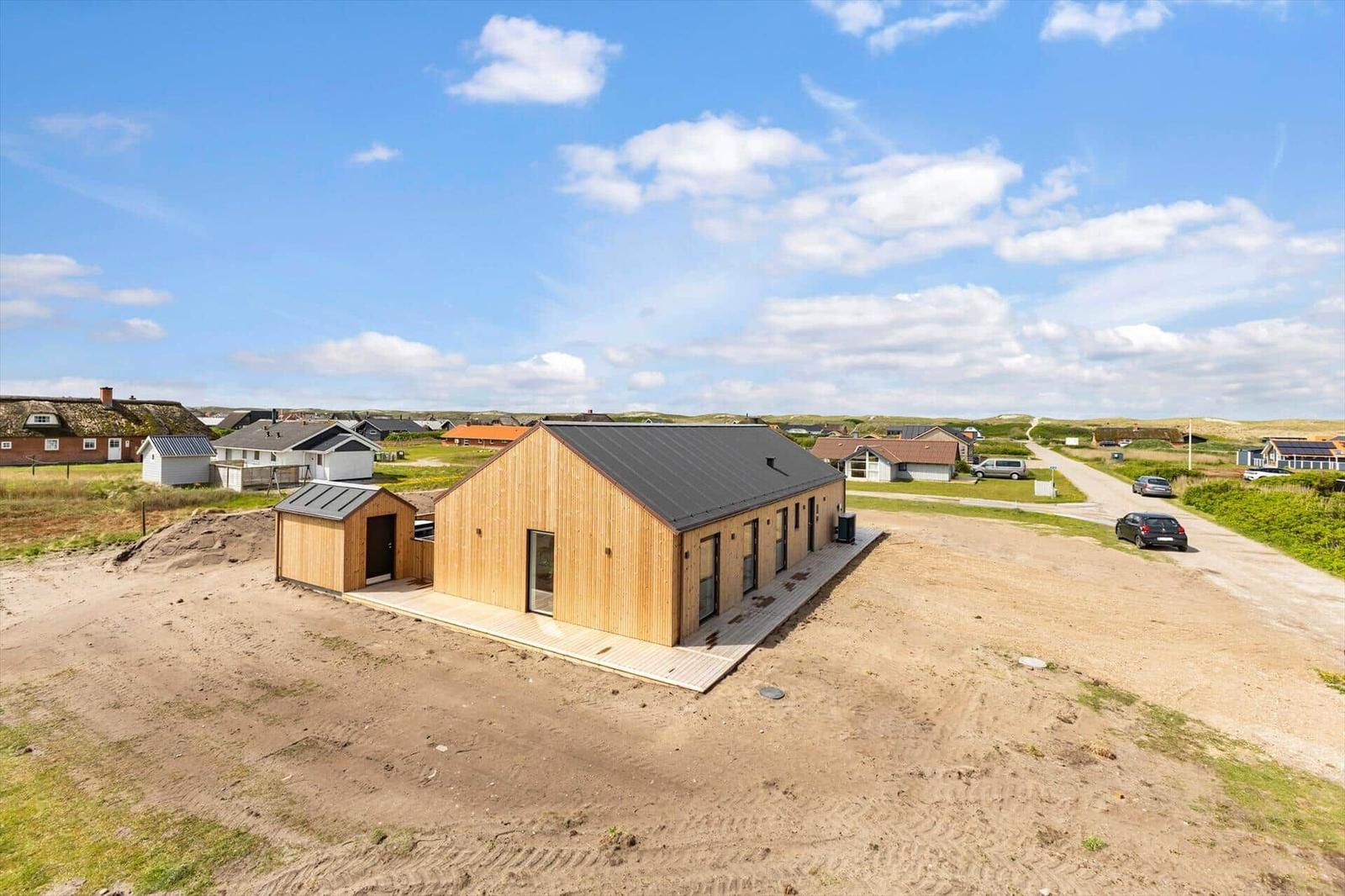 Moderne Holzhütte mit dunklem Dach auf sandigem Grundstück. Umgebung mit weiteren Häusern und Wegen.