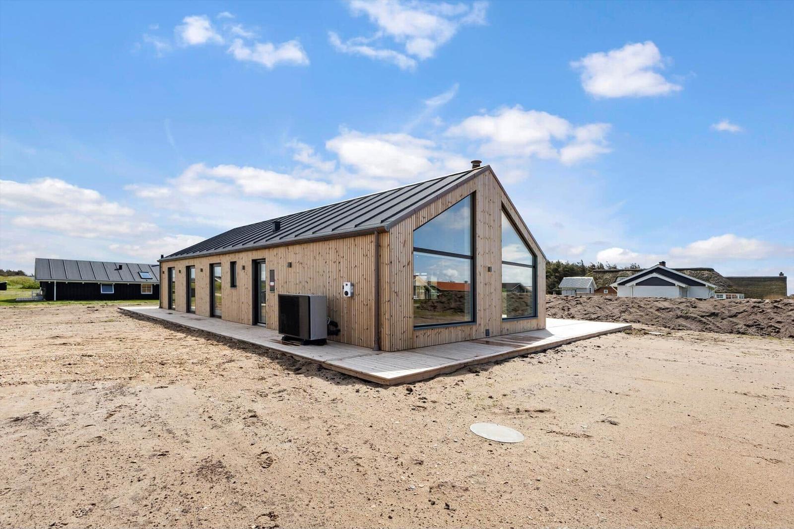 Moderne Holzhütte mit großem Fenster und dunklem Dach auf sandigem Grundstück.