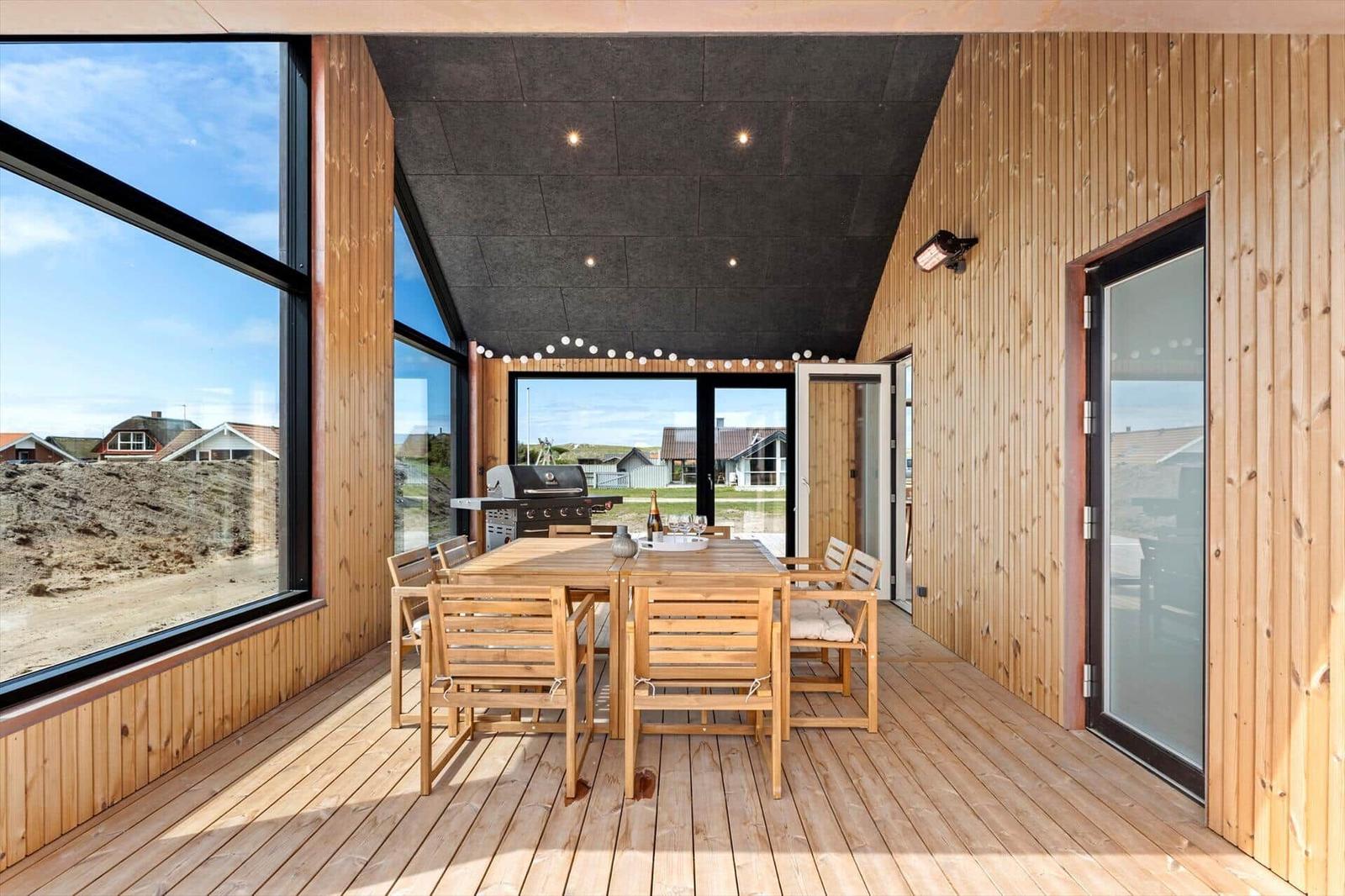 Terrasse mit Holztisch, Stühlen und Grill. Große Fenster mit Blick auf Strand.