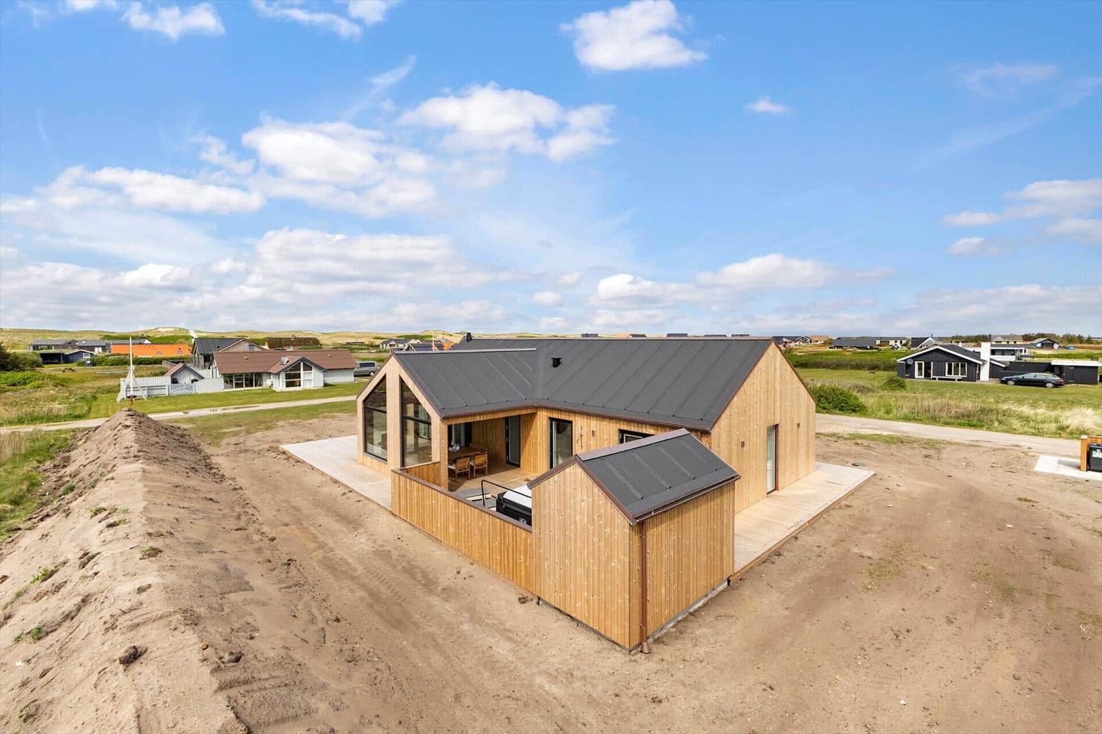 Moderne Holzhütte mit großem Balkon und dunklem Dach auf sandigem Grundstück.